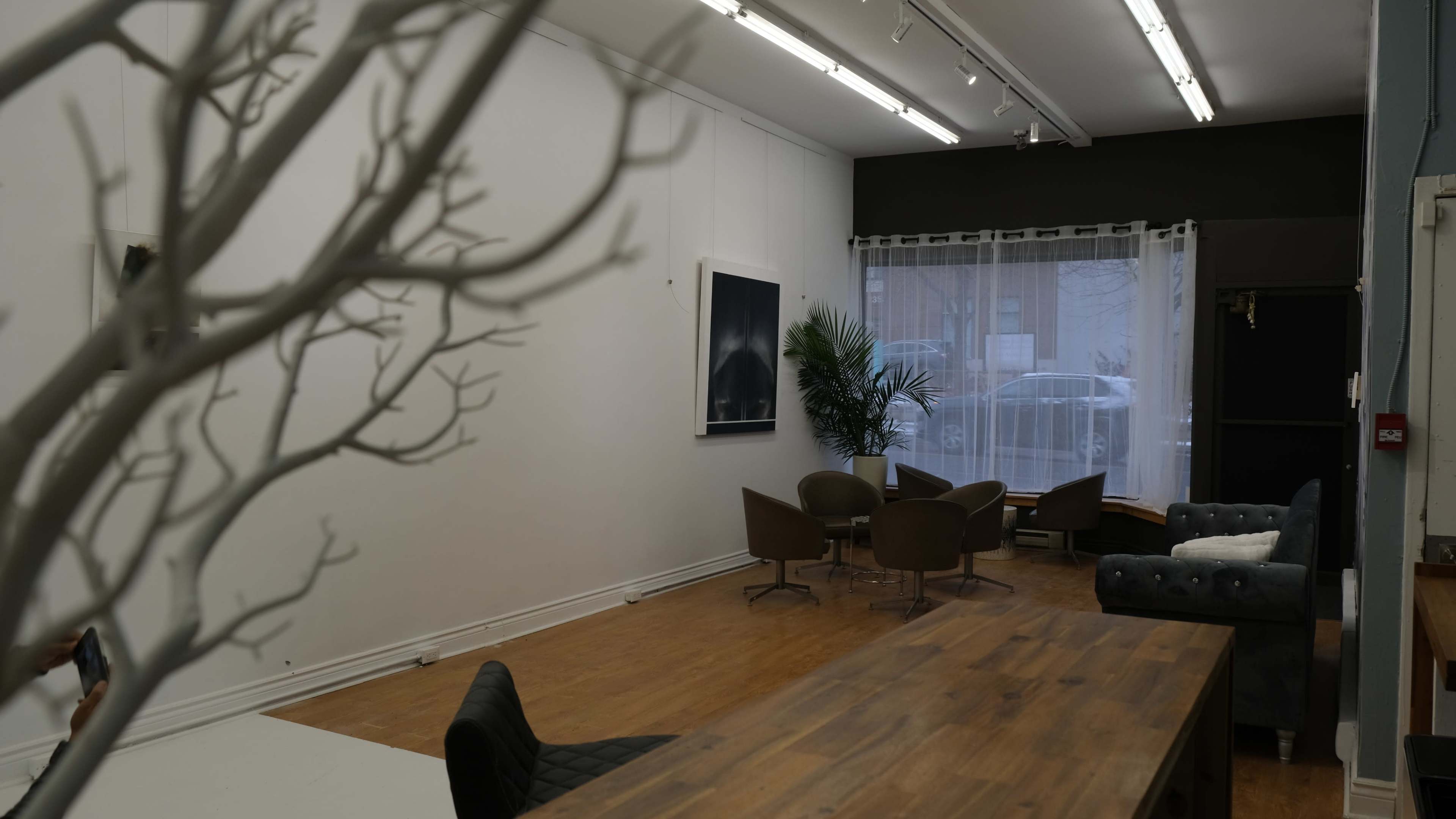 A spacious room with a large wooden table, several chairs, and a window with sheer curtains letting in natural light.