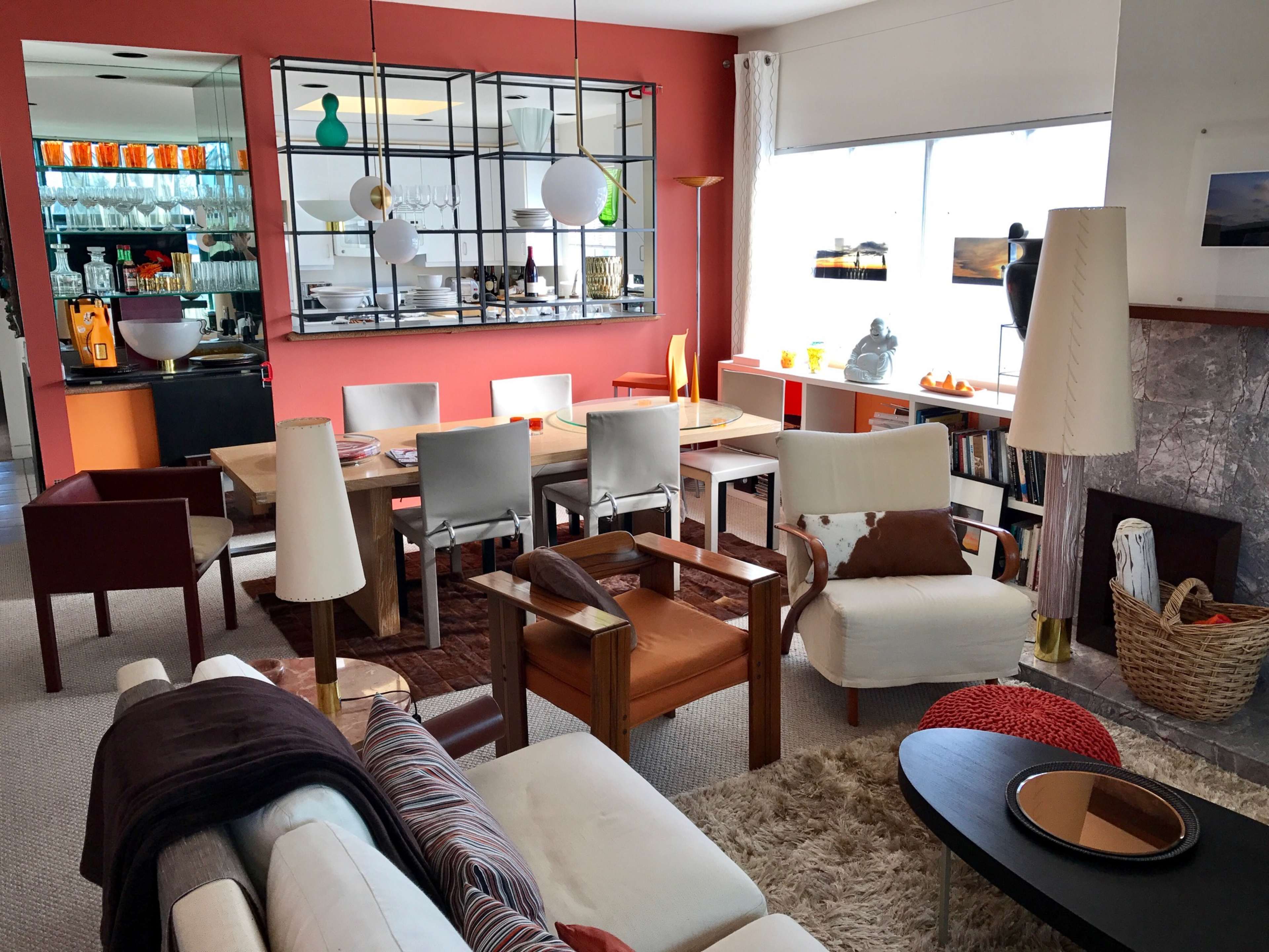 The image shows a modern living room and dining area with a mix of furniture including a sofa, armchairs, and a dining table, set against a backdrop of orange walls and a large window.