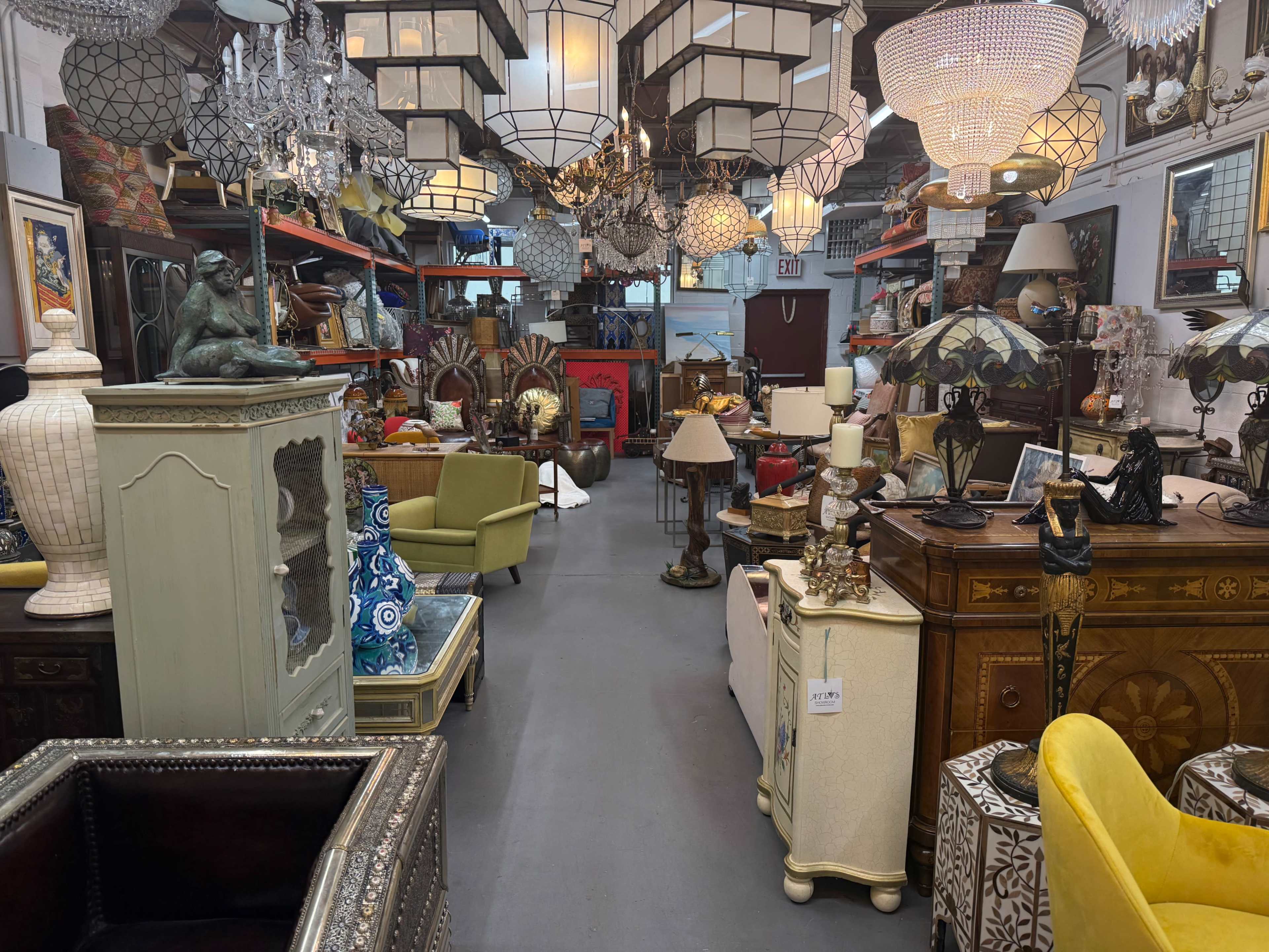 The image shows a spacious antique store filled with various decorative items, including furniture, lamps, and chandeliers, arranged along the aisles.