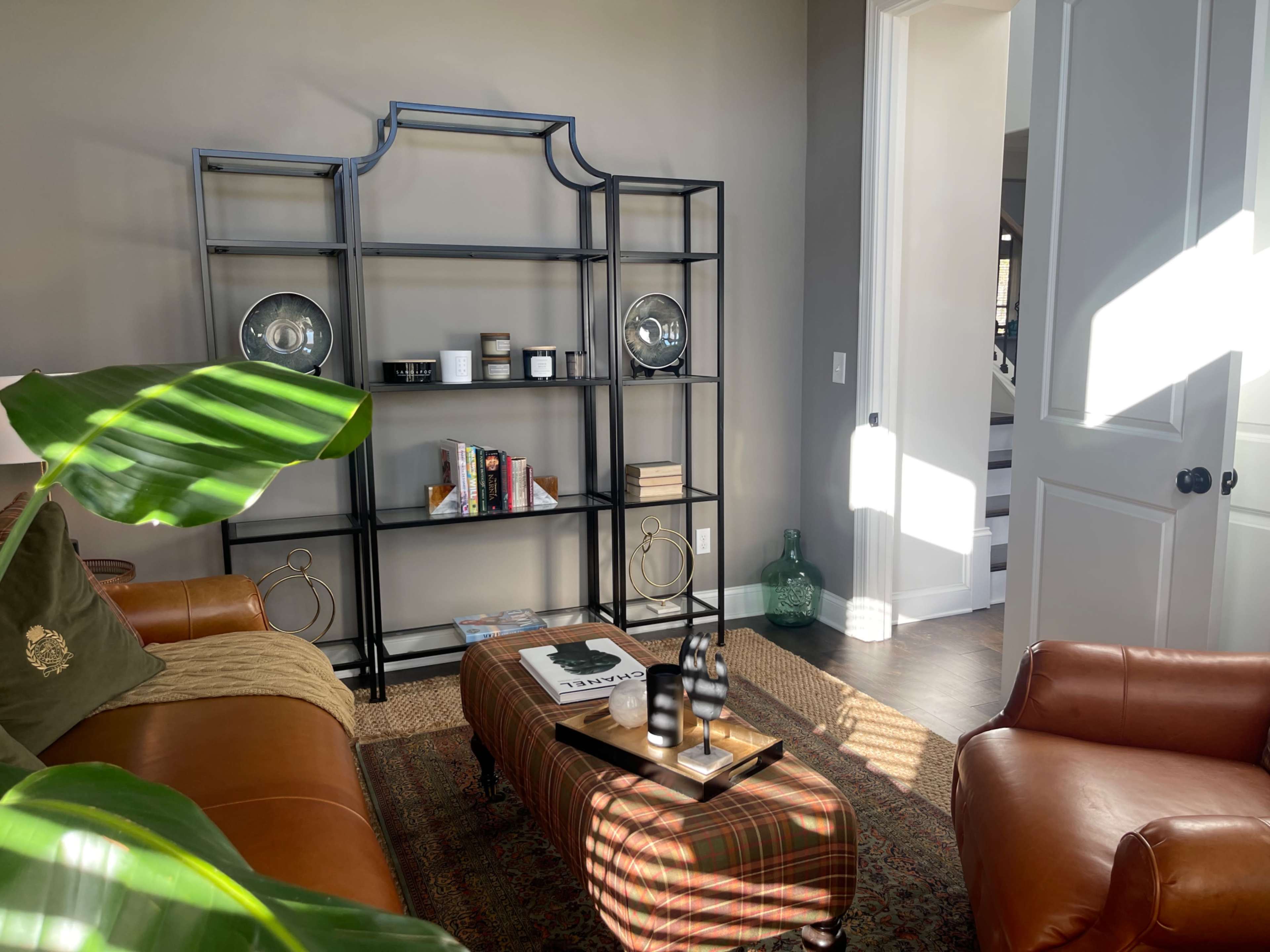 A modern living room features a black metal shelving unit, a brown leather ottoman, and a green plant, with sunlight illuminating the space.