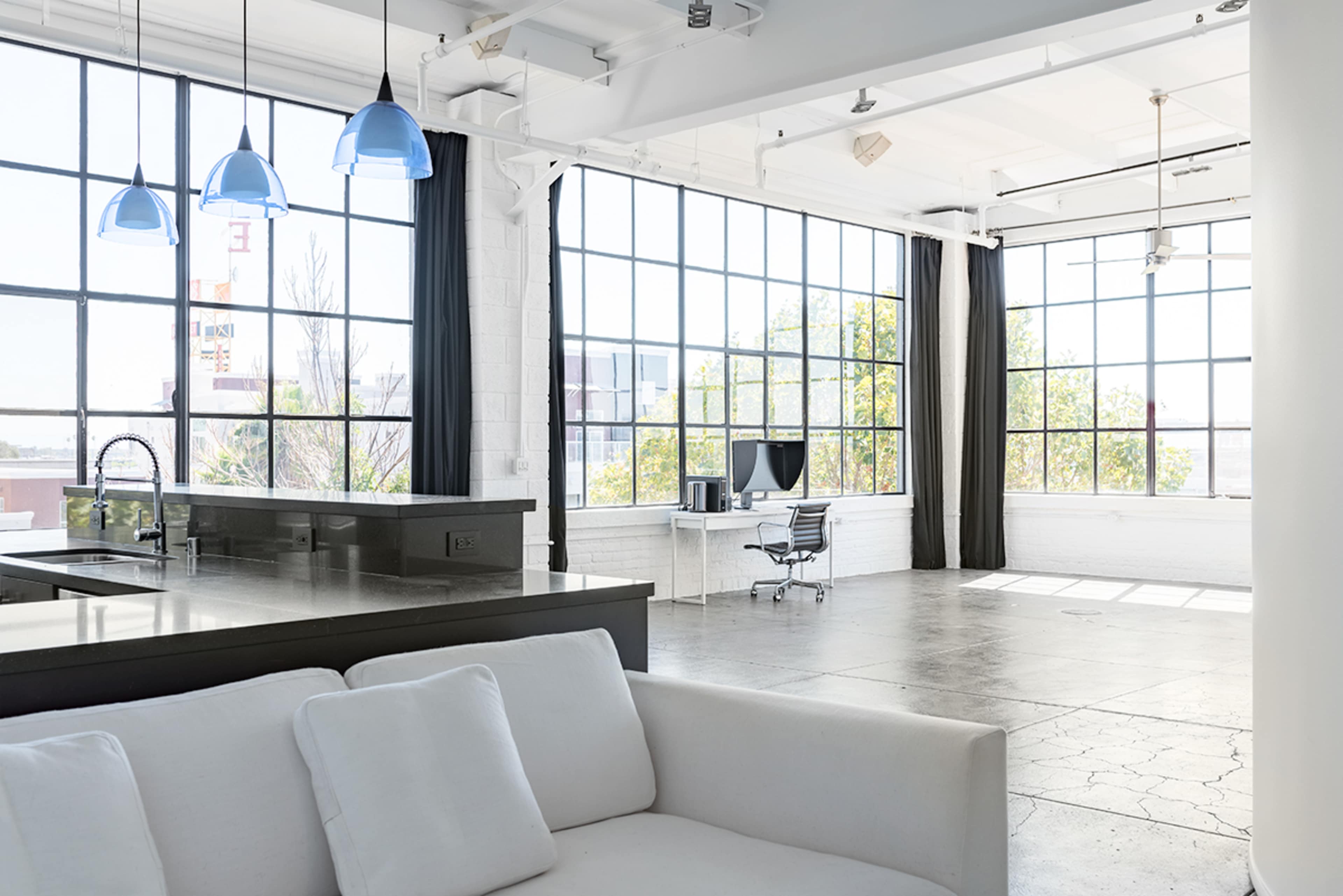 A spacious, modern loft features large windows, a minimalist kitchen island, and an organized work area with a desk and chair.