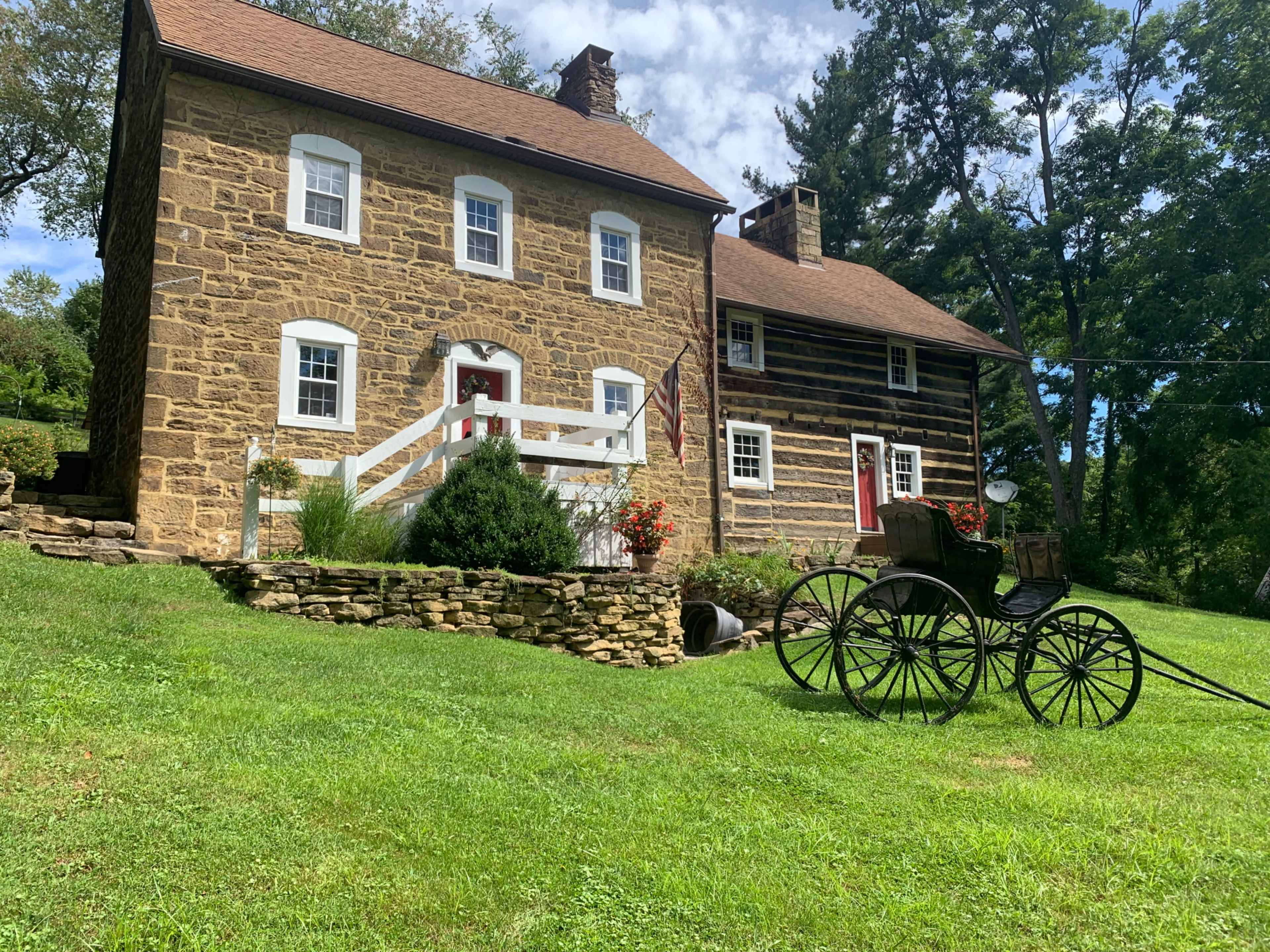 Two historic homes, one made of stone and the other of wood, with a grassy area and a vintage horse-drawn carriage in the foreground.