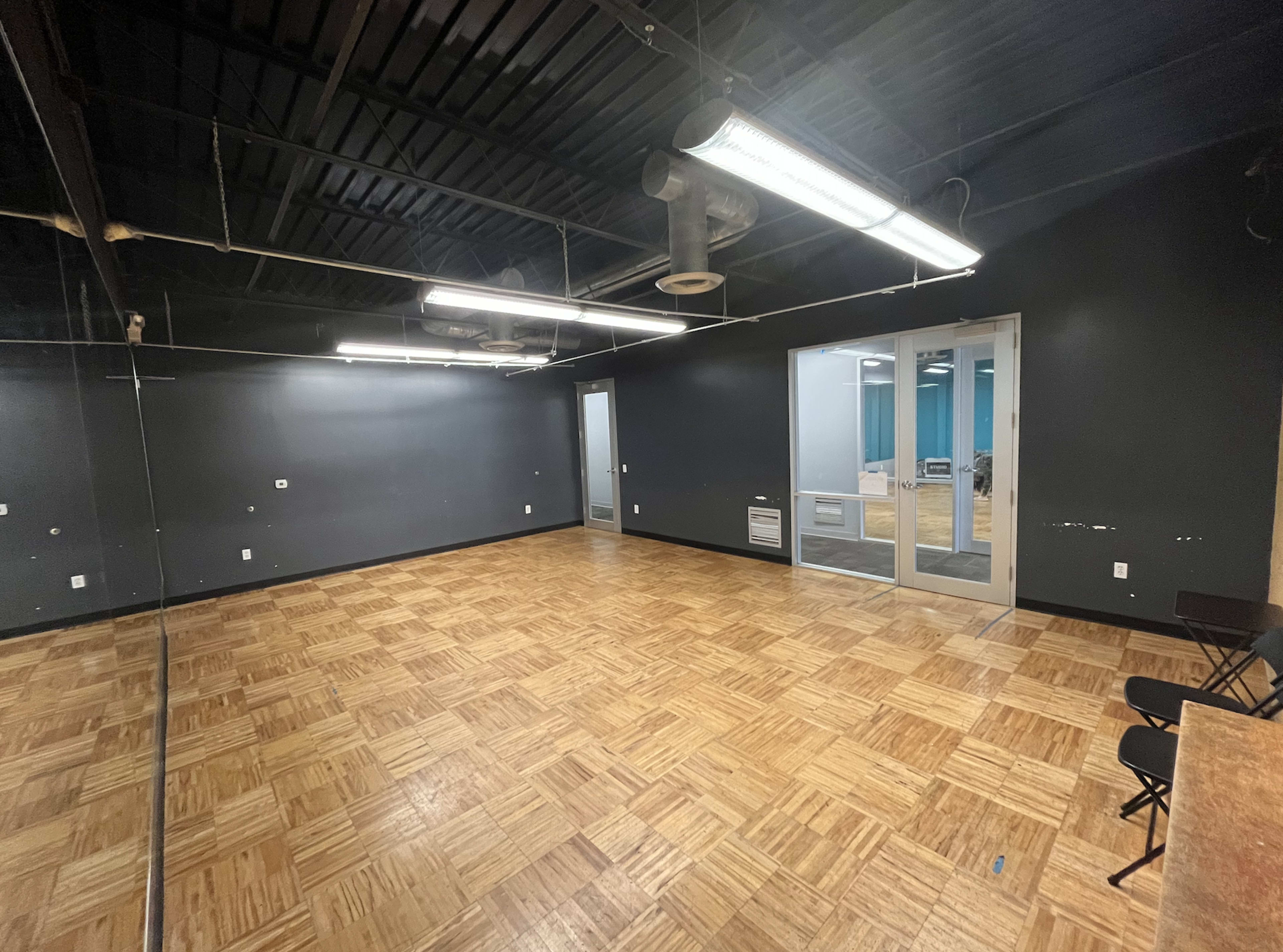 The image shows a spacious room with dark walls, a wooden floor, and a large mirror on one side, featuring a glass door that leads to an adjacent area.