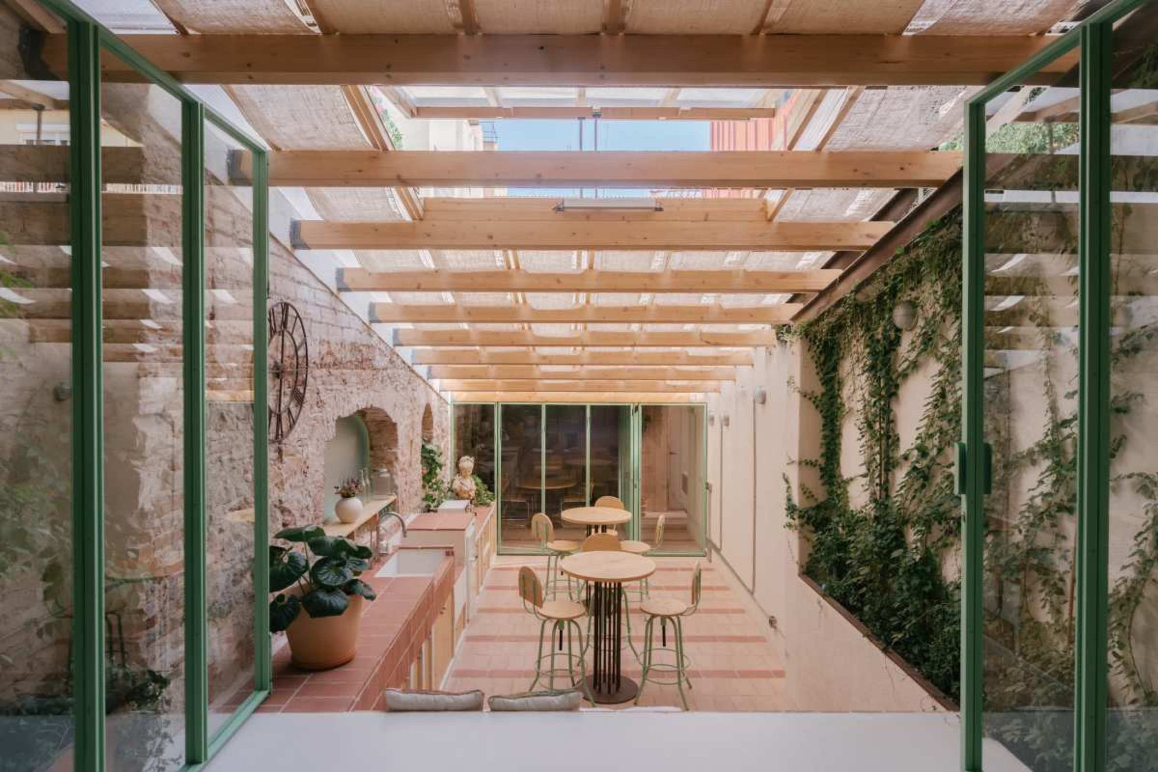 The image shows a light-filled courtyard with wooden framing, green framed windows, and potted plants, featuring two round tables and four chairs.