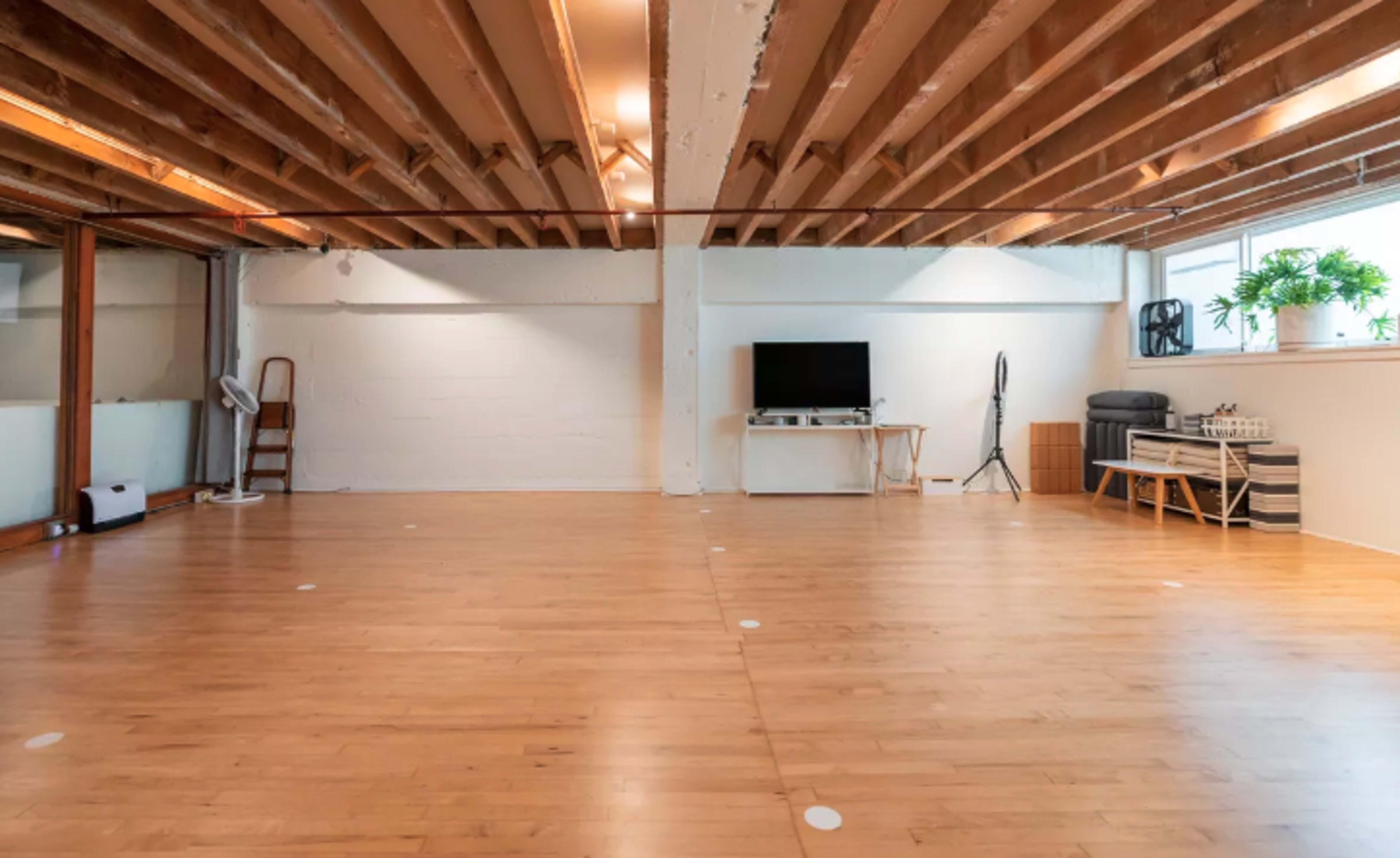 A spacious room with wooden flooring, exposed beam ceilings, and minimalist furnishings including a television and a chair.
