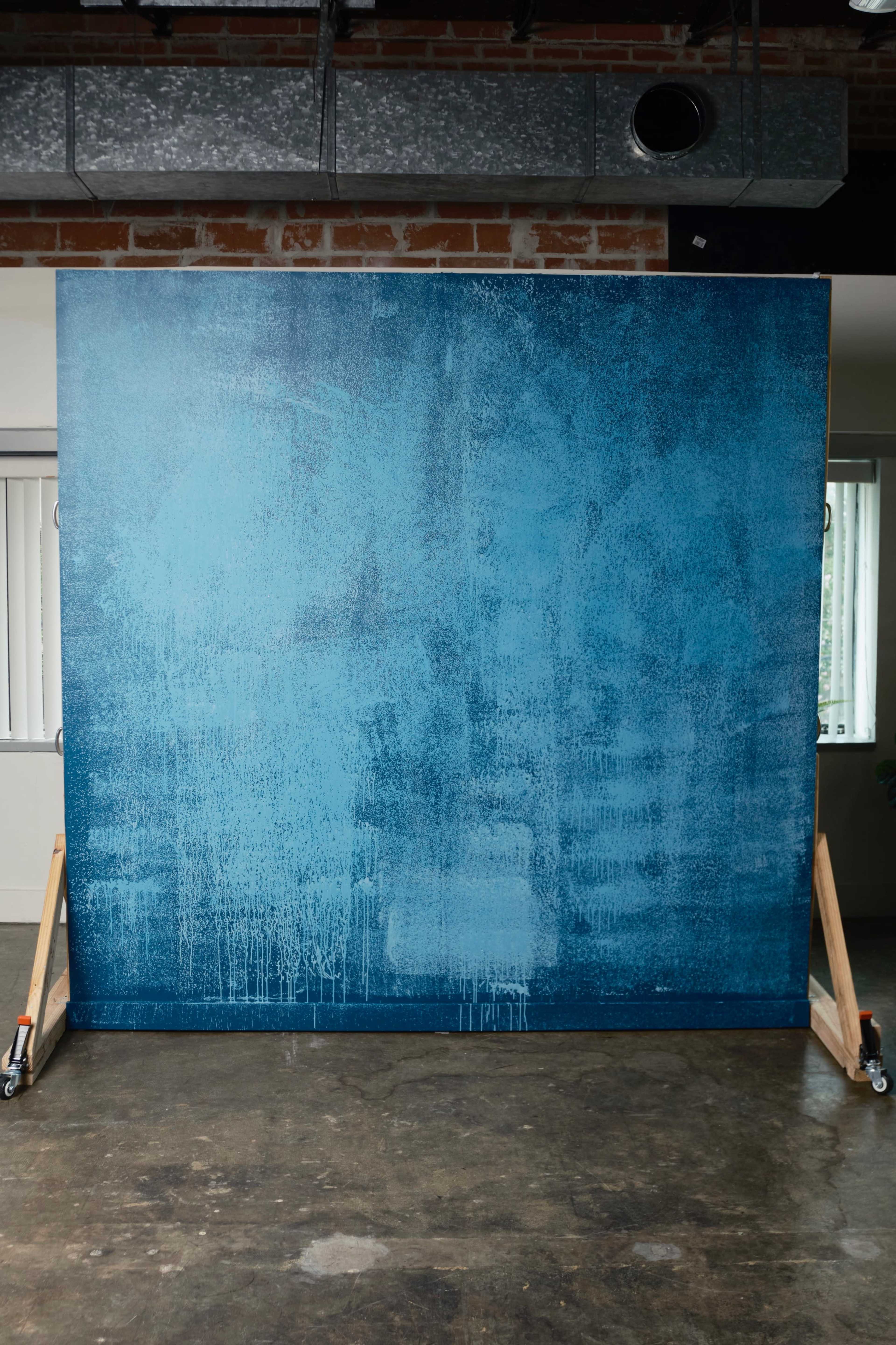 The image shows a large blue backdrop mounted on a wooden frame with wheels, set against a brick wall in a spacious interior.