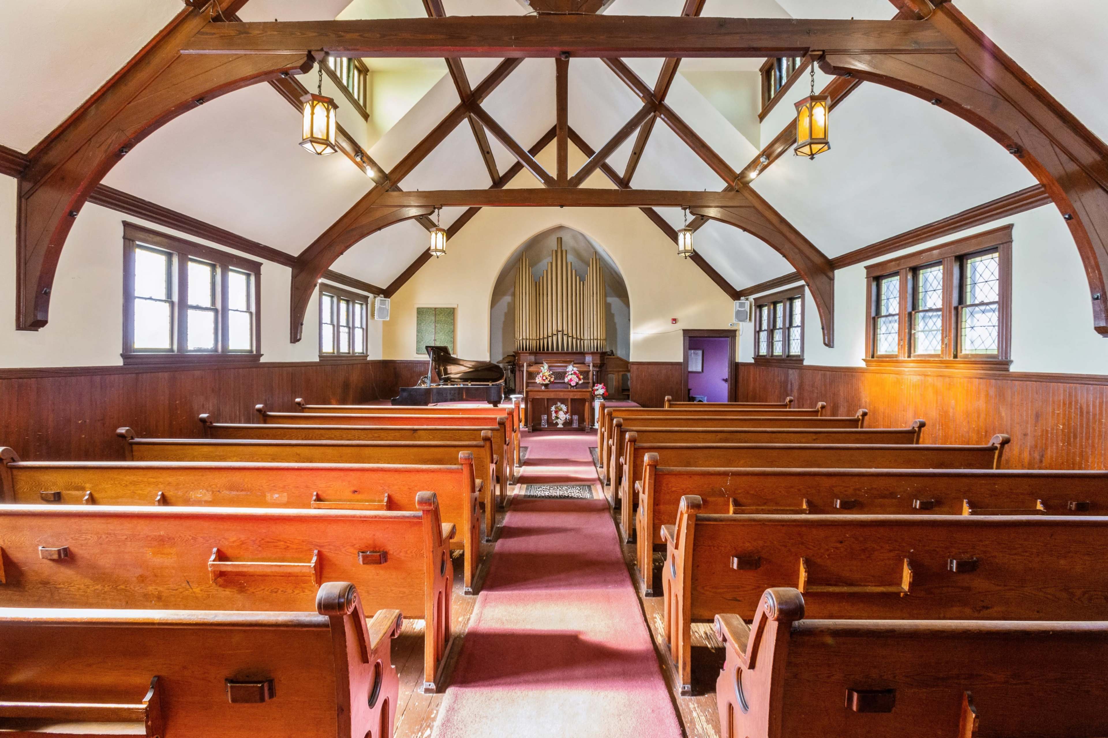 The interior of a church features wooden pews arranged in rows facing a piano and an organ at the front, with exposed beams and large windows.
