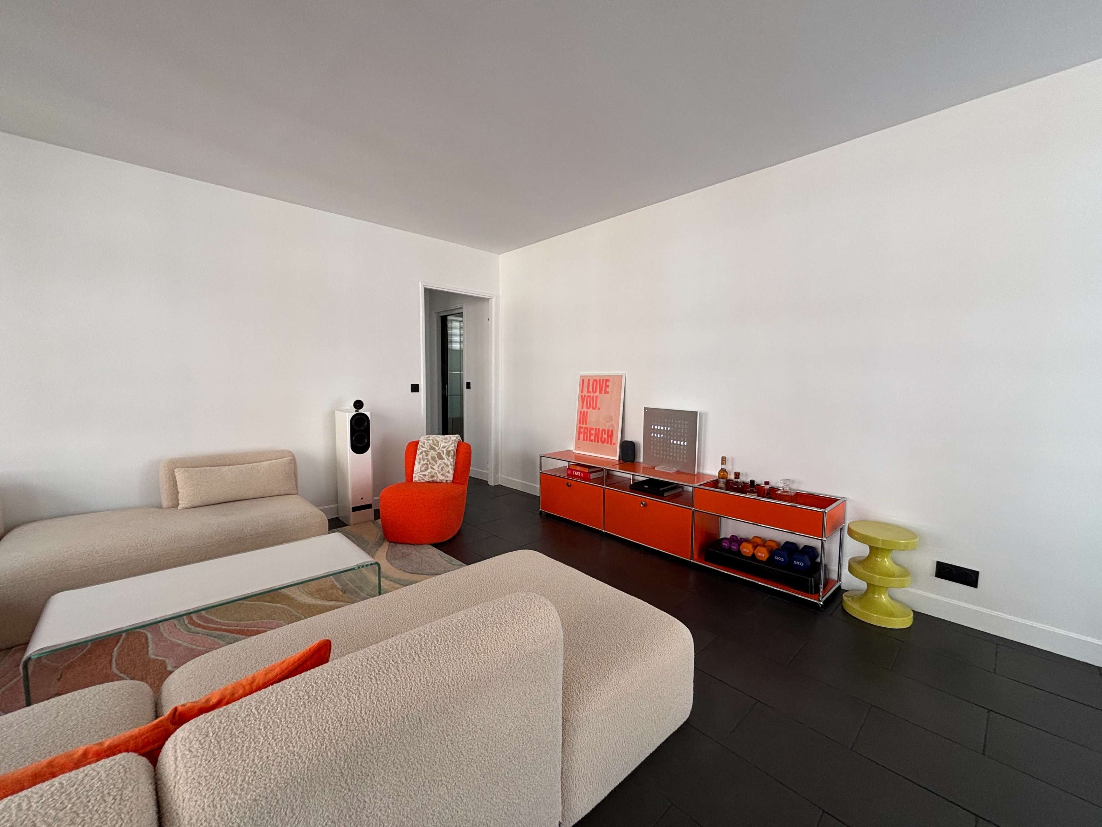 The living room features a minimalist design with two light-colored armchairs, a red accent chair, a television stand with bright decor, and a green side table.