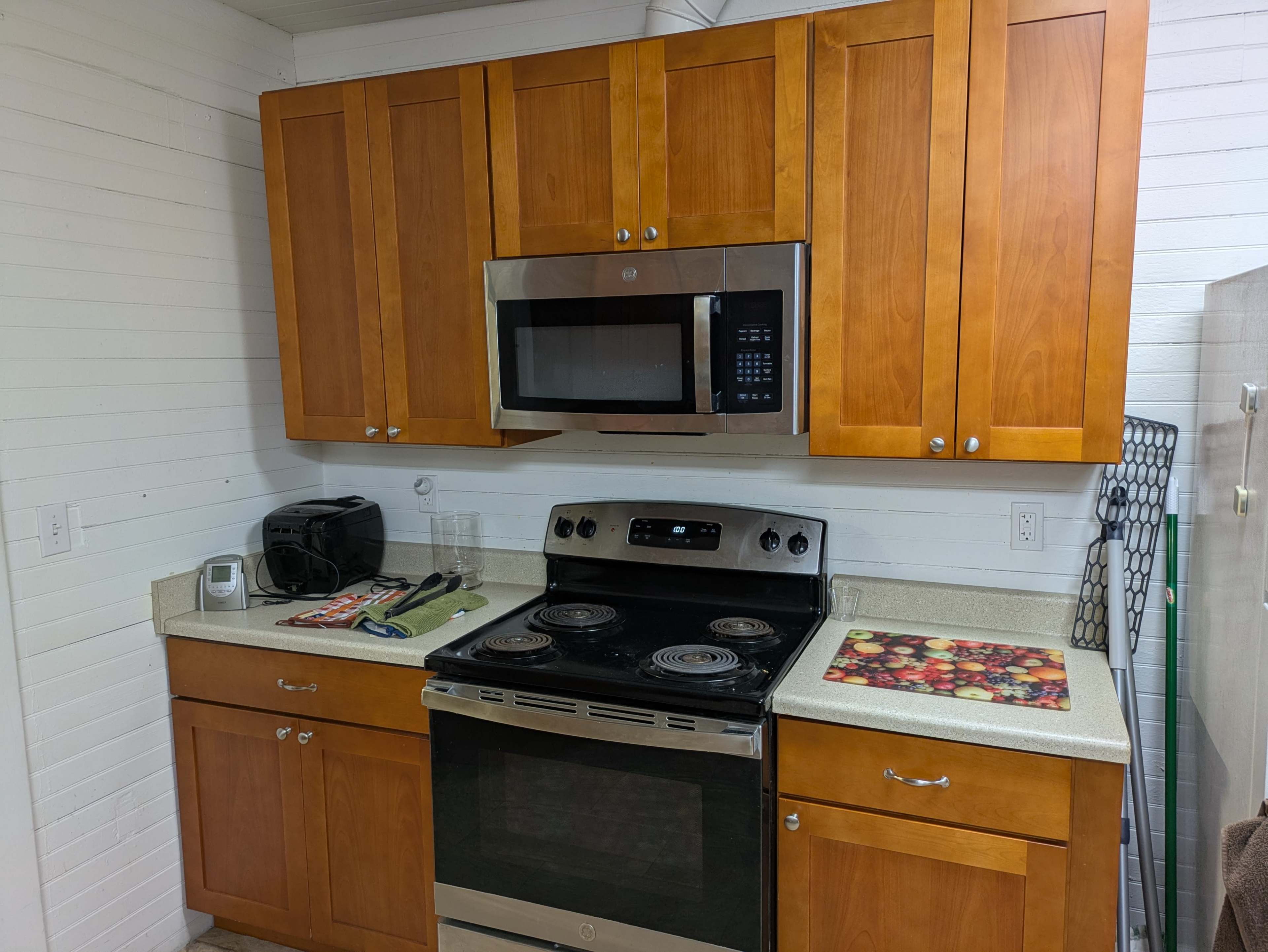 The image shows a kitchen with wooden cabinets, a stainless steel microwave above a black stove with four burners, and a countertop displaying a toaster and colorful cutting boards.