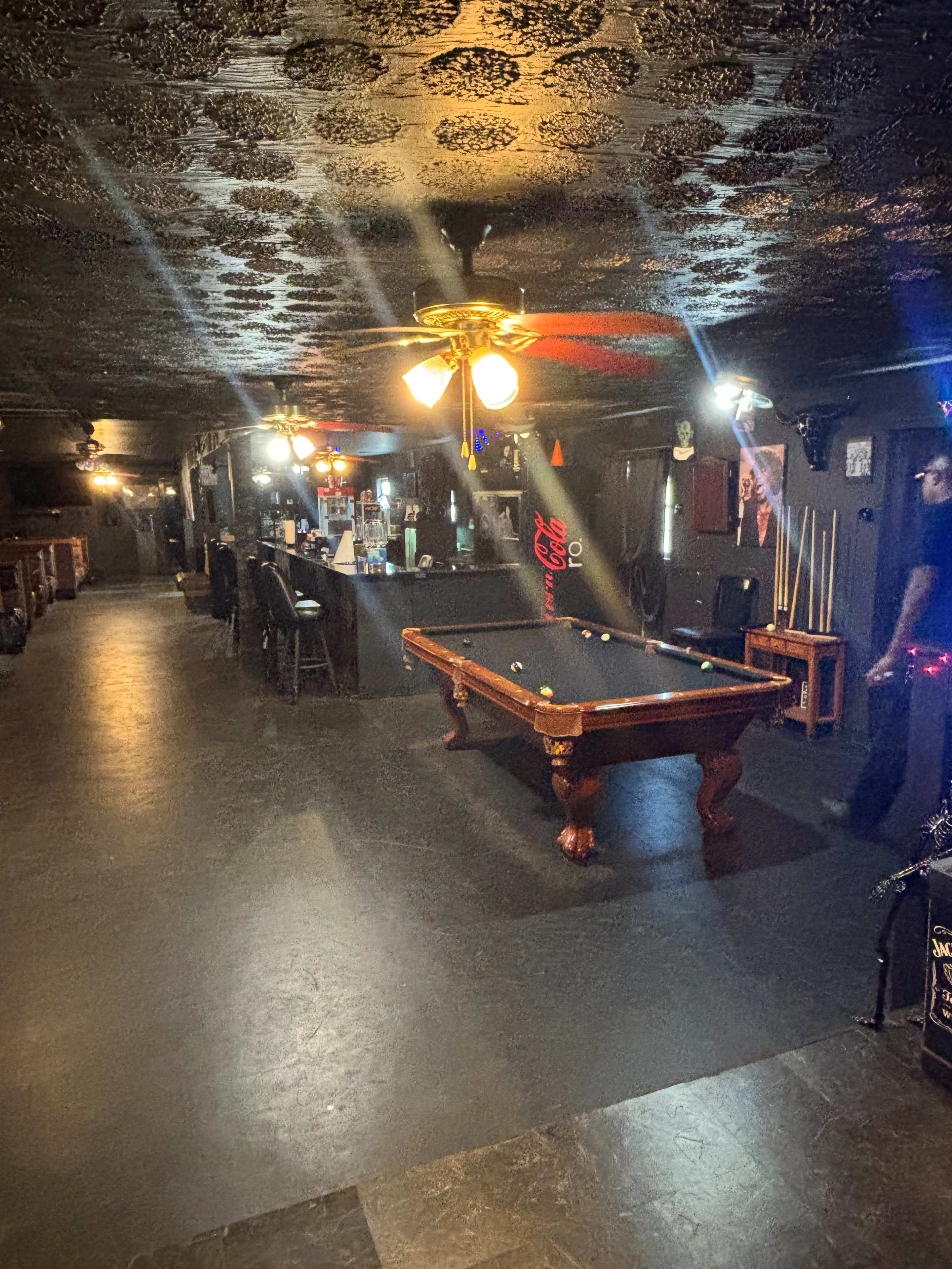 A dimly lit room features a billiard table surrounded by chairs and bar stools, with a ceiling fan overhead.