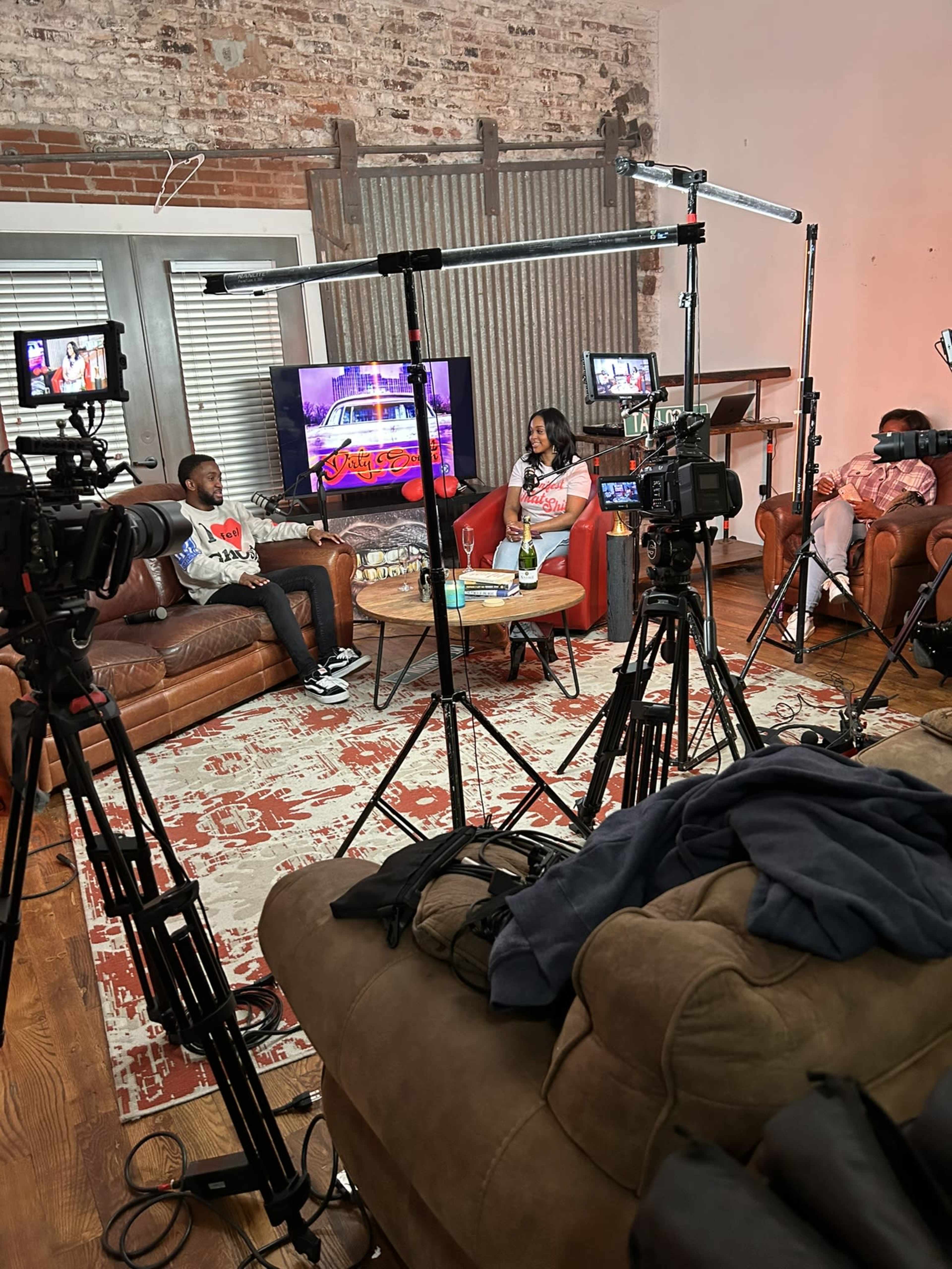 A television studio setup with cameras and lights surrounding two people seated on a couch, discussing on a set featuring a colorful backdrop.