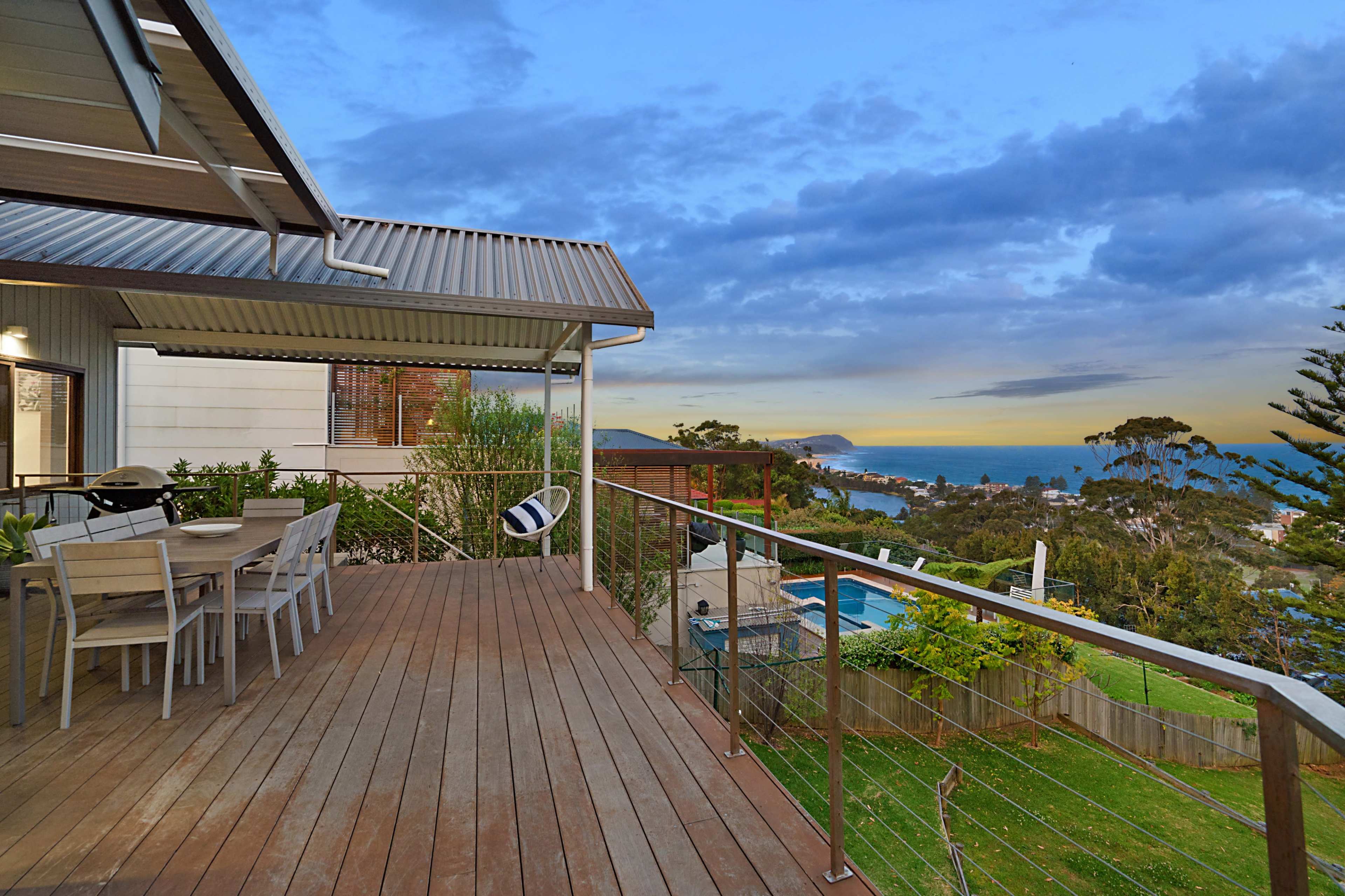 A wooden deck with a dining table overlooks a coastal view at sunset, featuring a swimming pool and surrounding greenery.