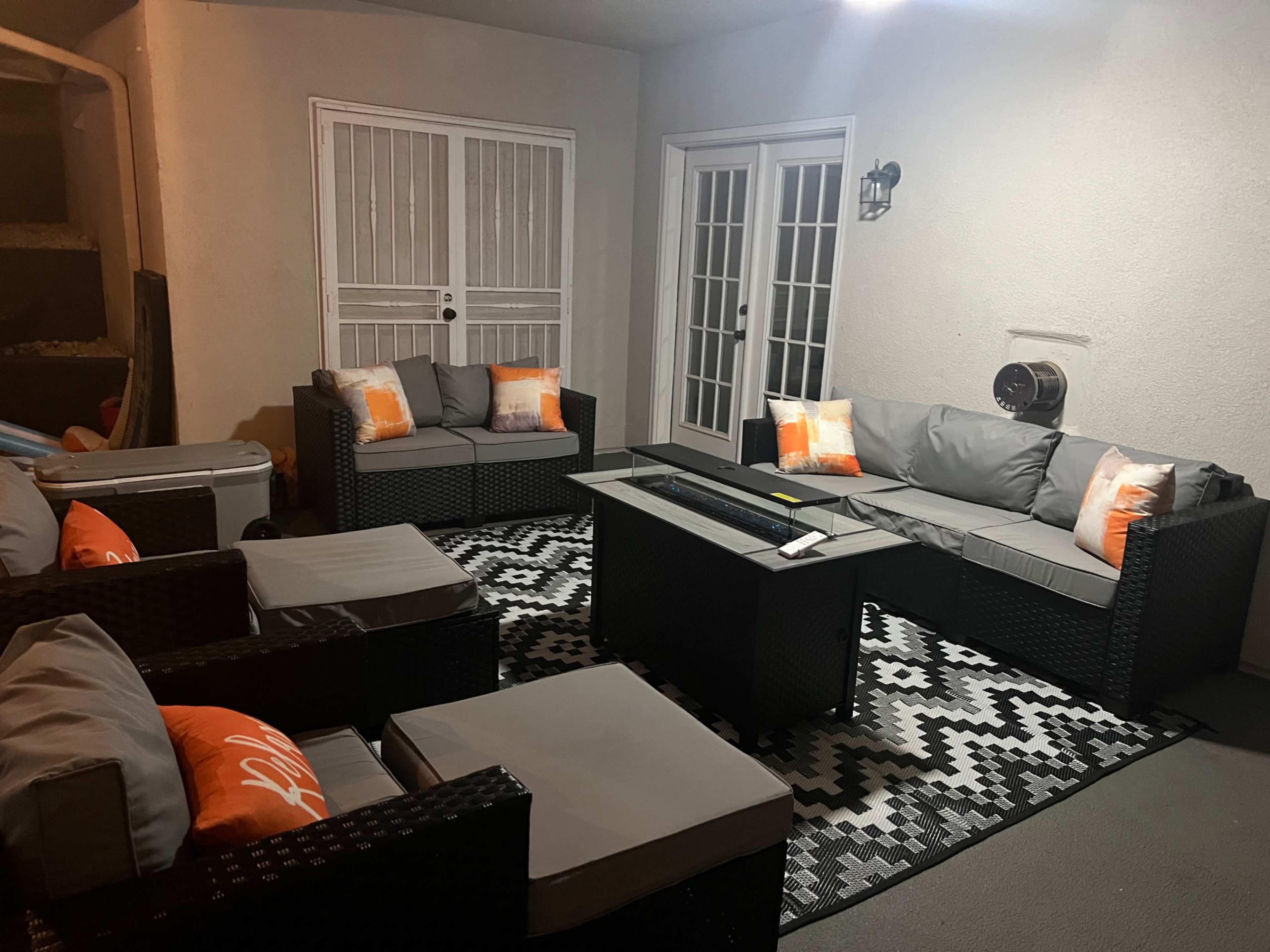 The image shows a patio area furnished with a sectional sofa, chairs, and a coffee table on a patterned rug, illuminated by indoor lighting.