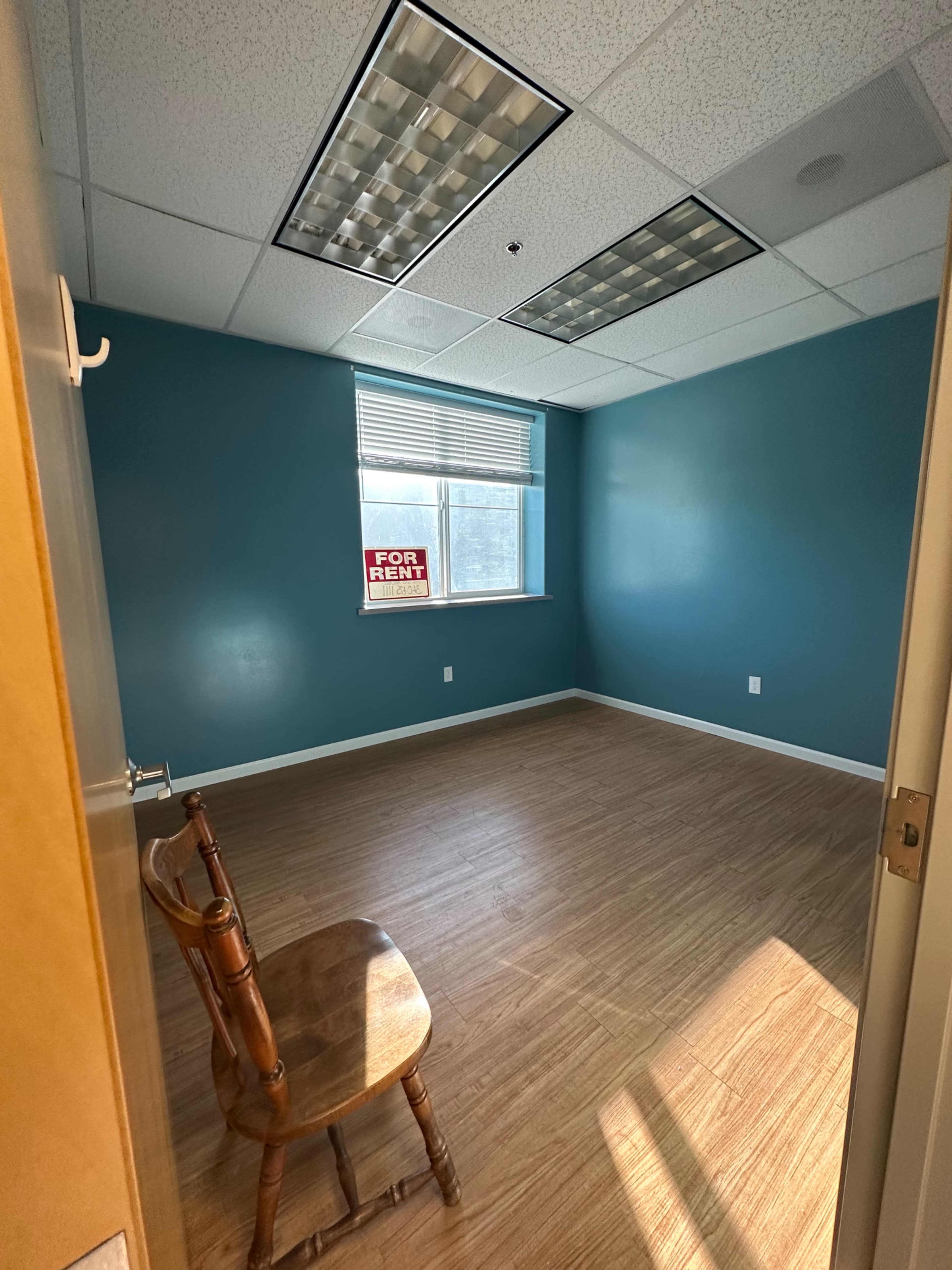 The image shows an empty room with teal walls, a lone wooden chair, and a "For Rent" sign visible in the window.