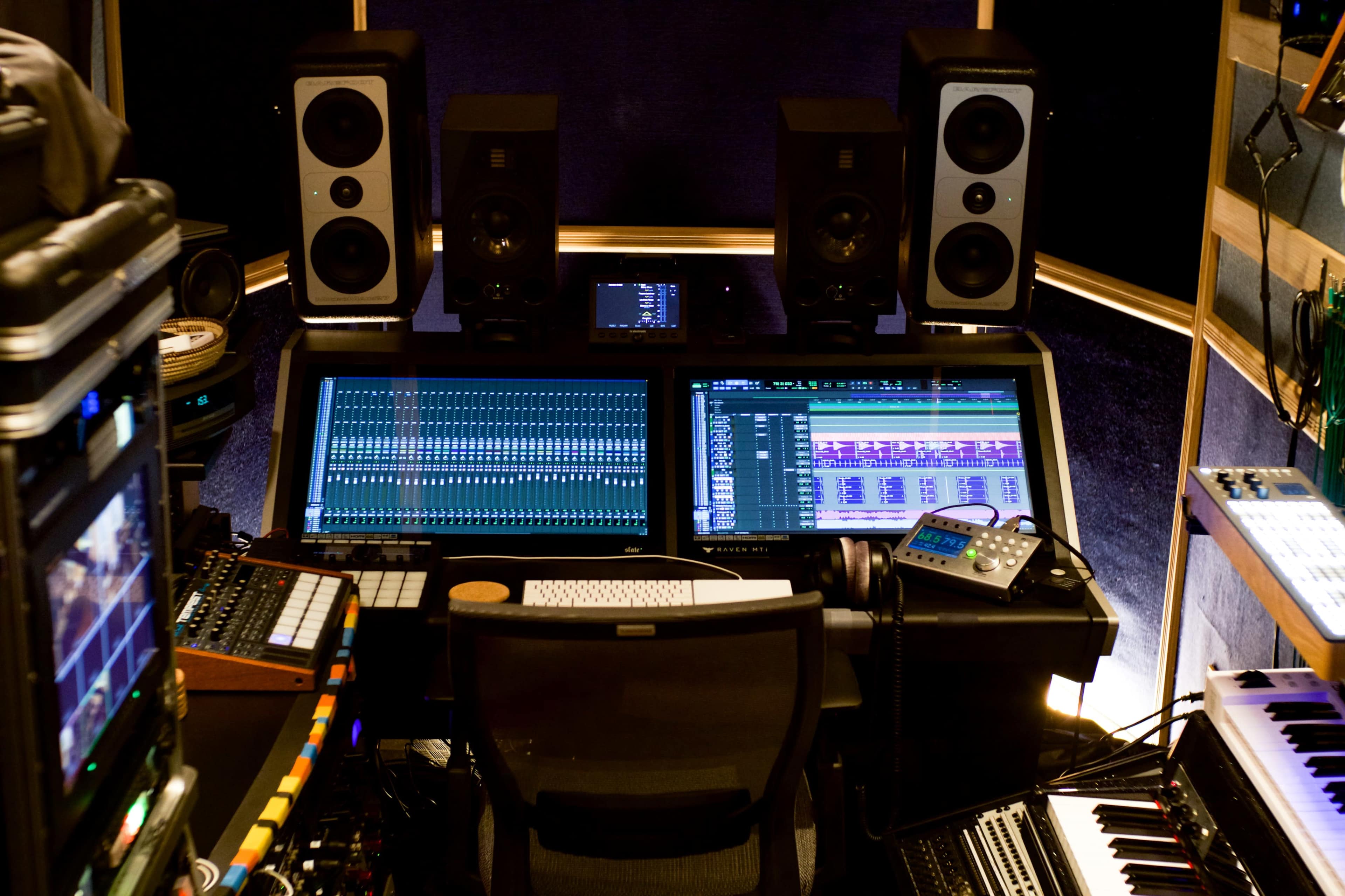 The image shows a music recording studio with two large computer monitors displaying audio editing software, surrounded by various instruments and audio equipment.