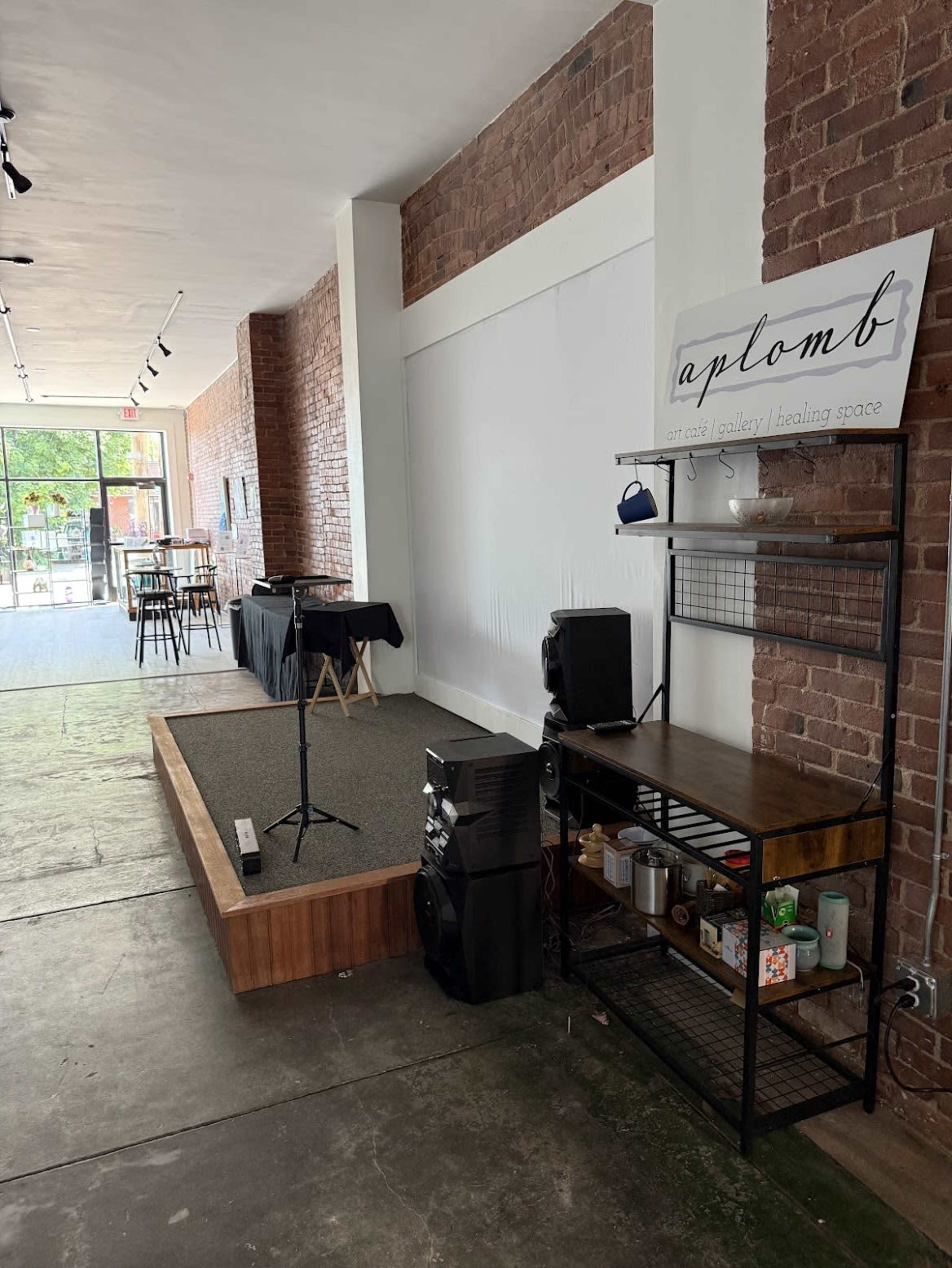 The image depicts an interior space featuring a small stage with a microphone, a sound system on a black shelf, and a sign that reads "aplomb," all within a brick-walled venue.