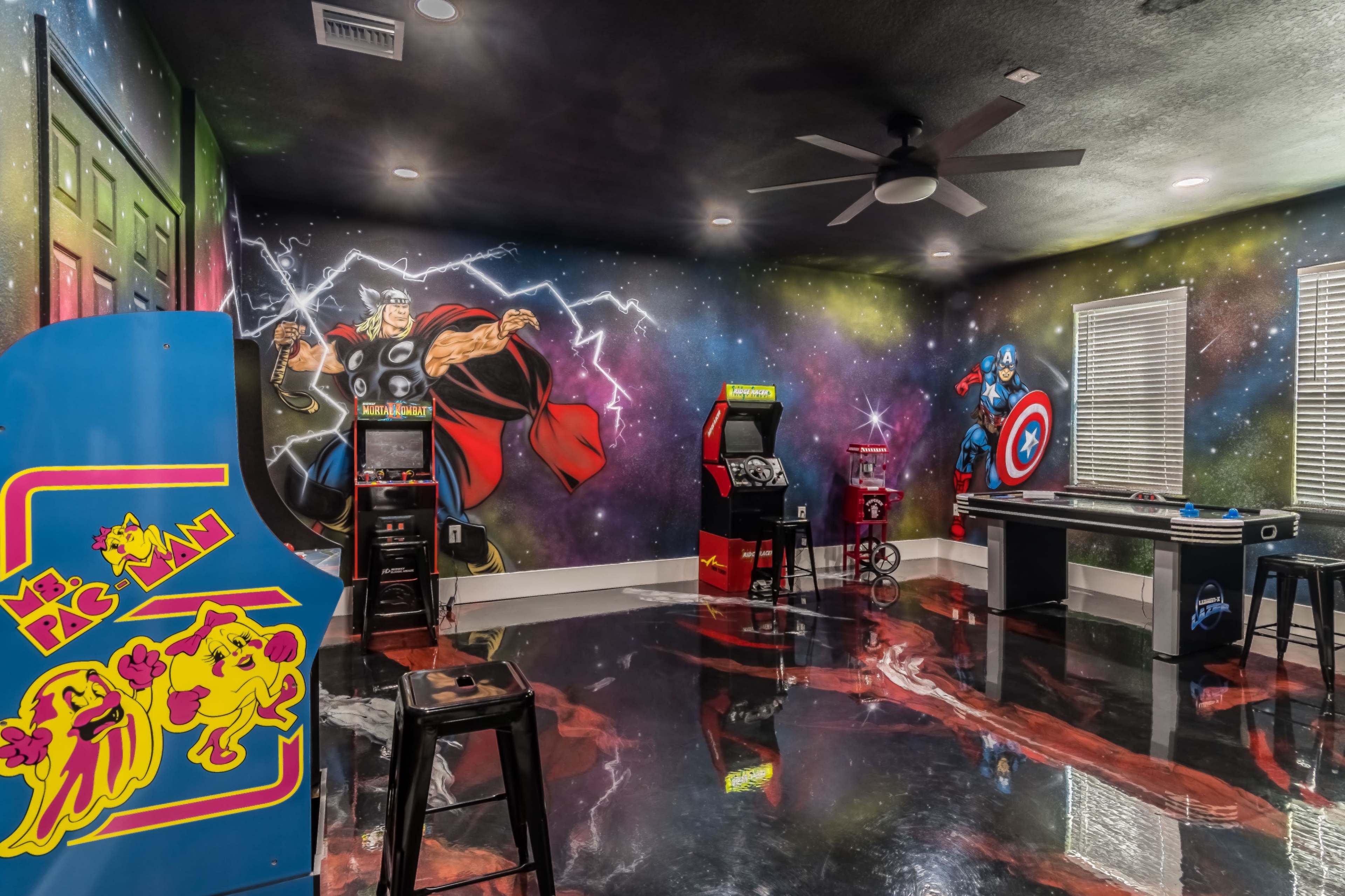 A colorful game room featuring superhero murals, an arcade cabinet, and various game tables against a dark floor.