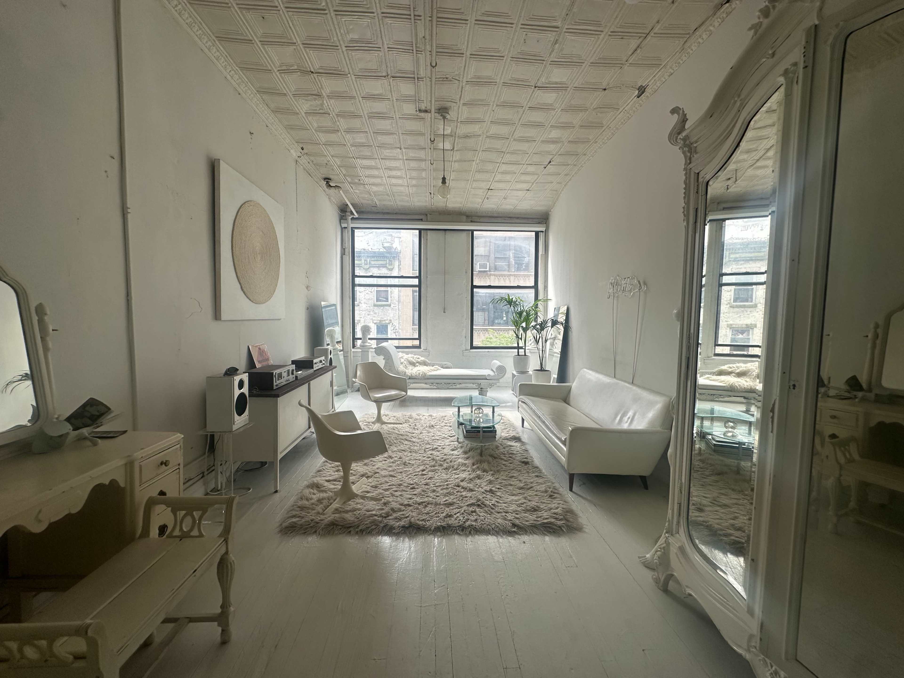 The image shows a bright, spacious room with large windows, featuring a white sofa, a glass coffee table, a decorative rug, and a full-length mirror.