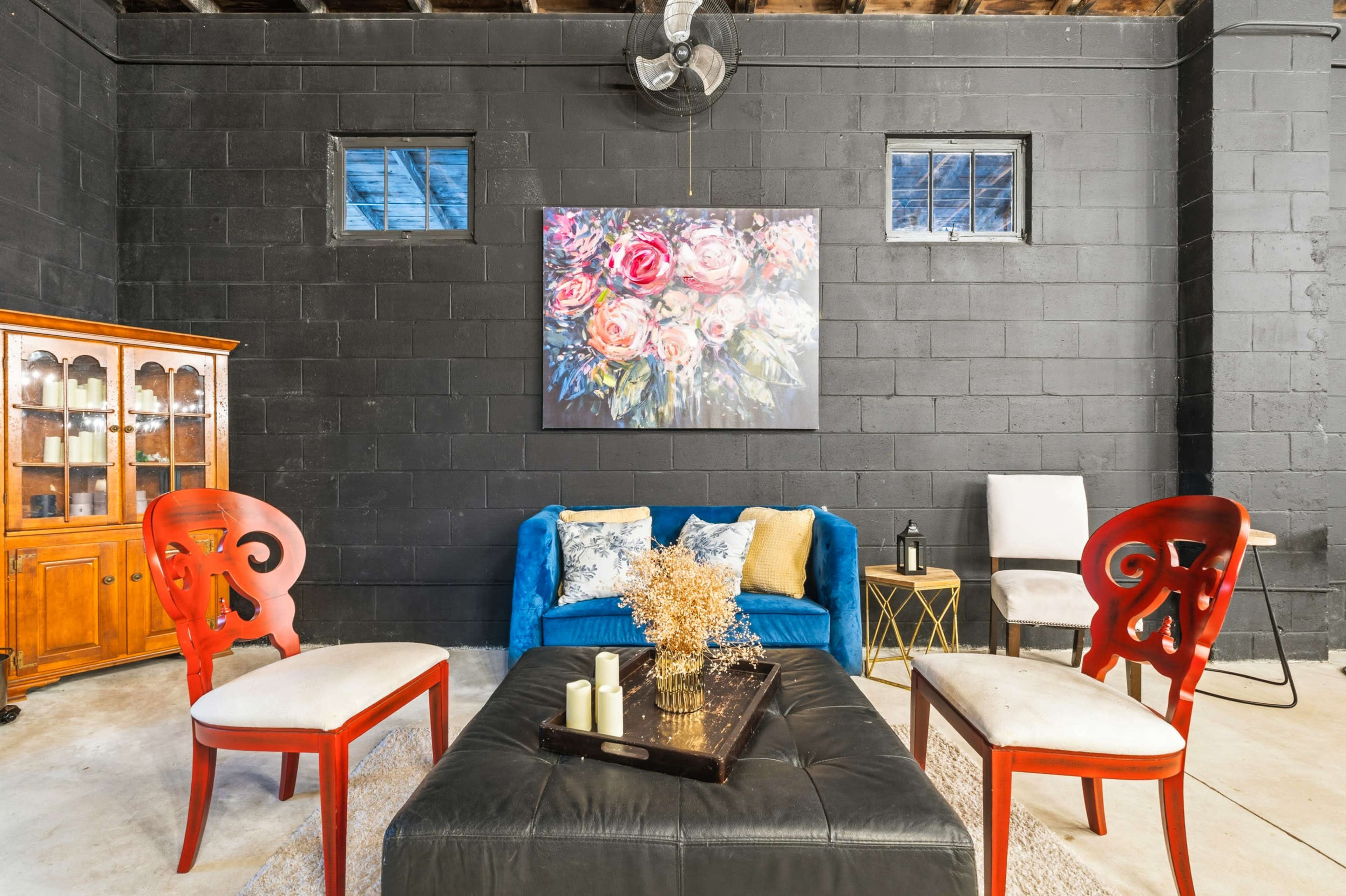 The image depicts a modern living space with a blue couch, two red chairs, and a black coffee table, set against a textured black wall adorned with a floral painting.