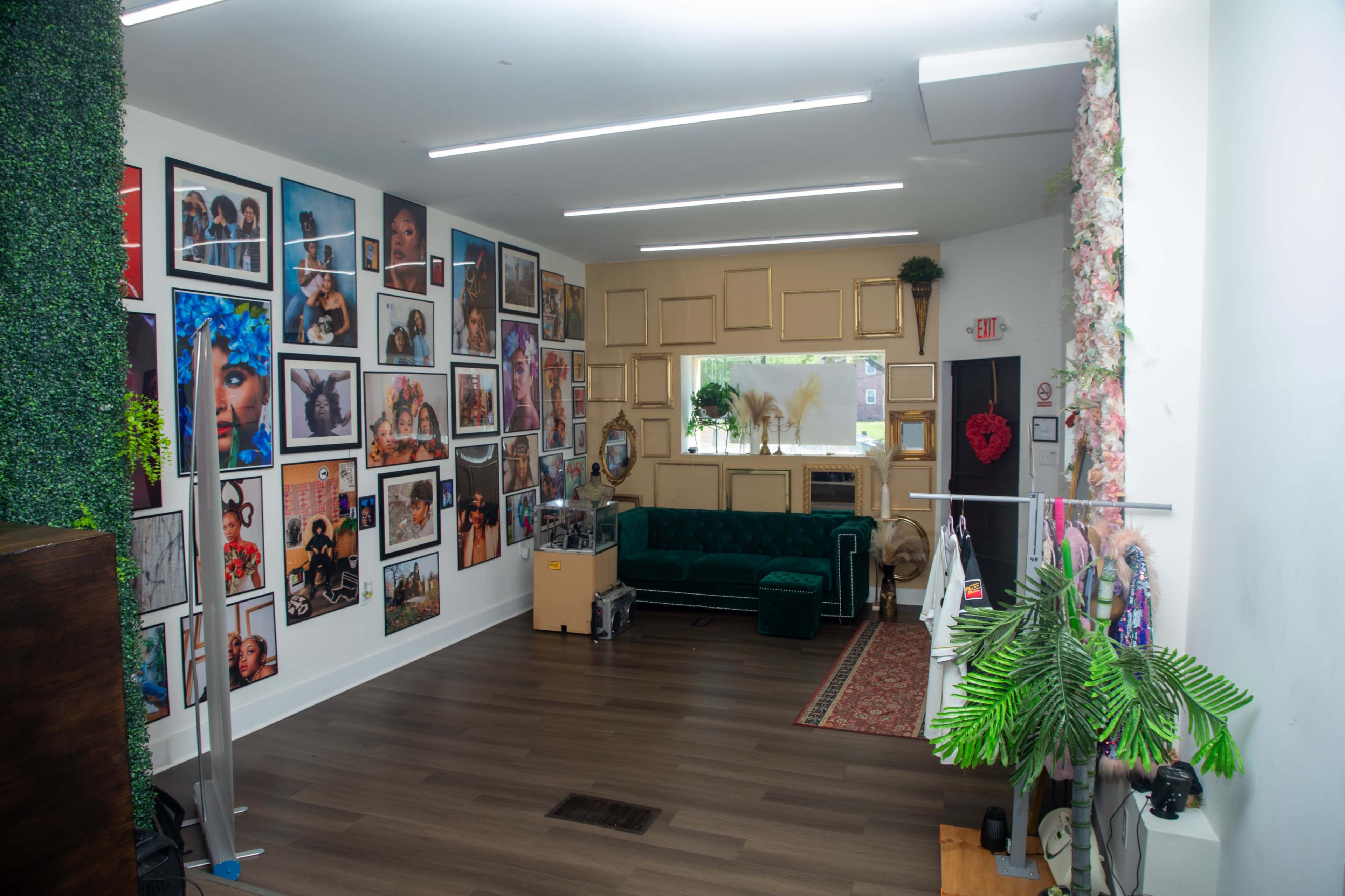 The image shows a well-decorated studio or salon space with a green velvet sofa, a collection of photo frames on the walls, and plants placed throughout the room.