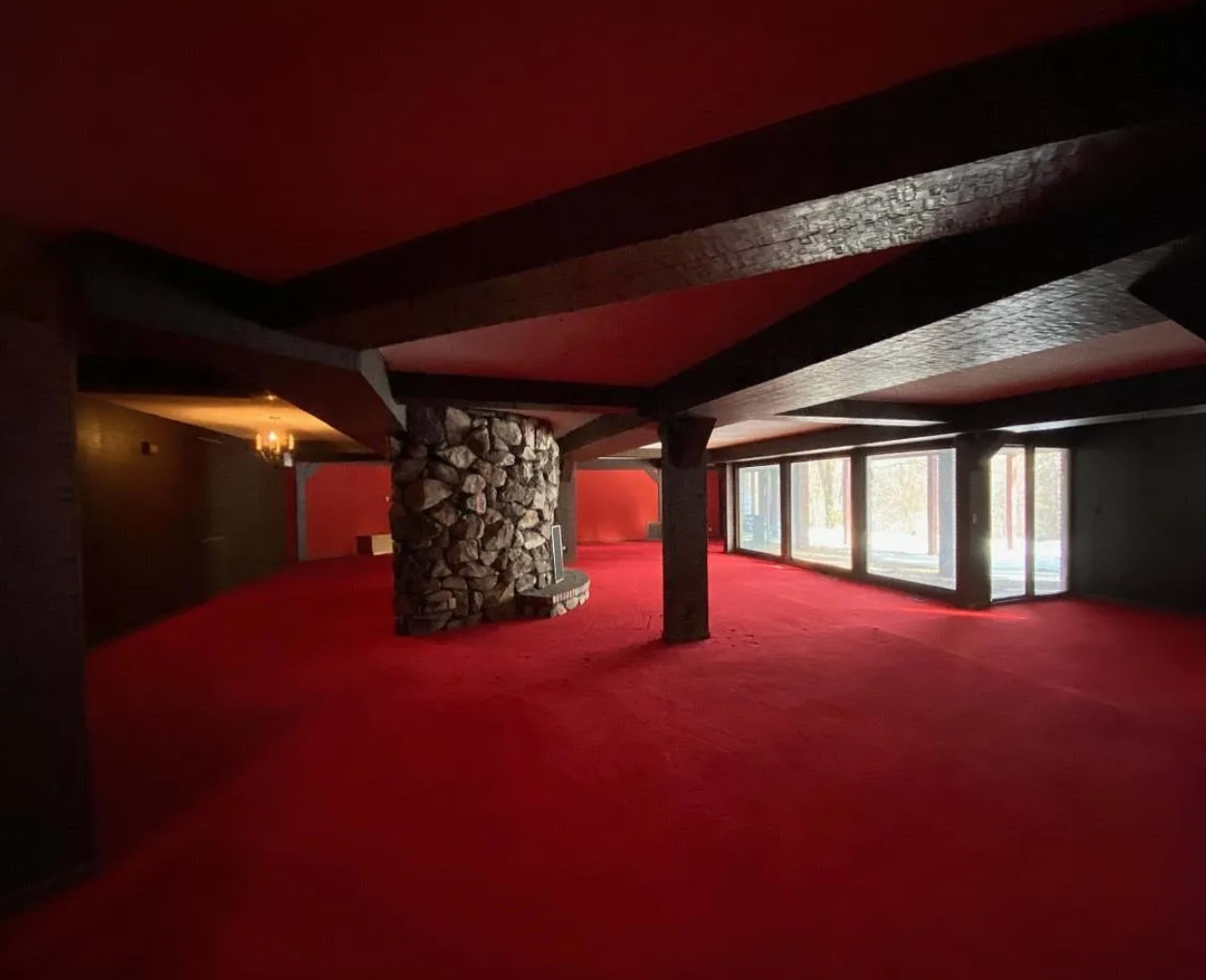 The image shows a large room with red carpet flooring, a stone fireplace, and large windows allowing natural light.