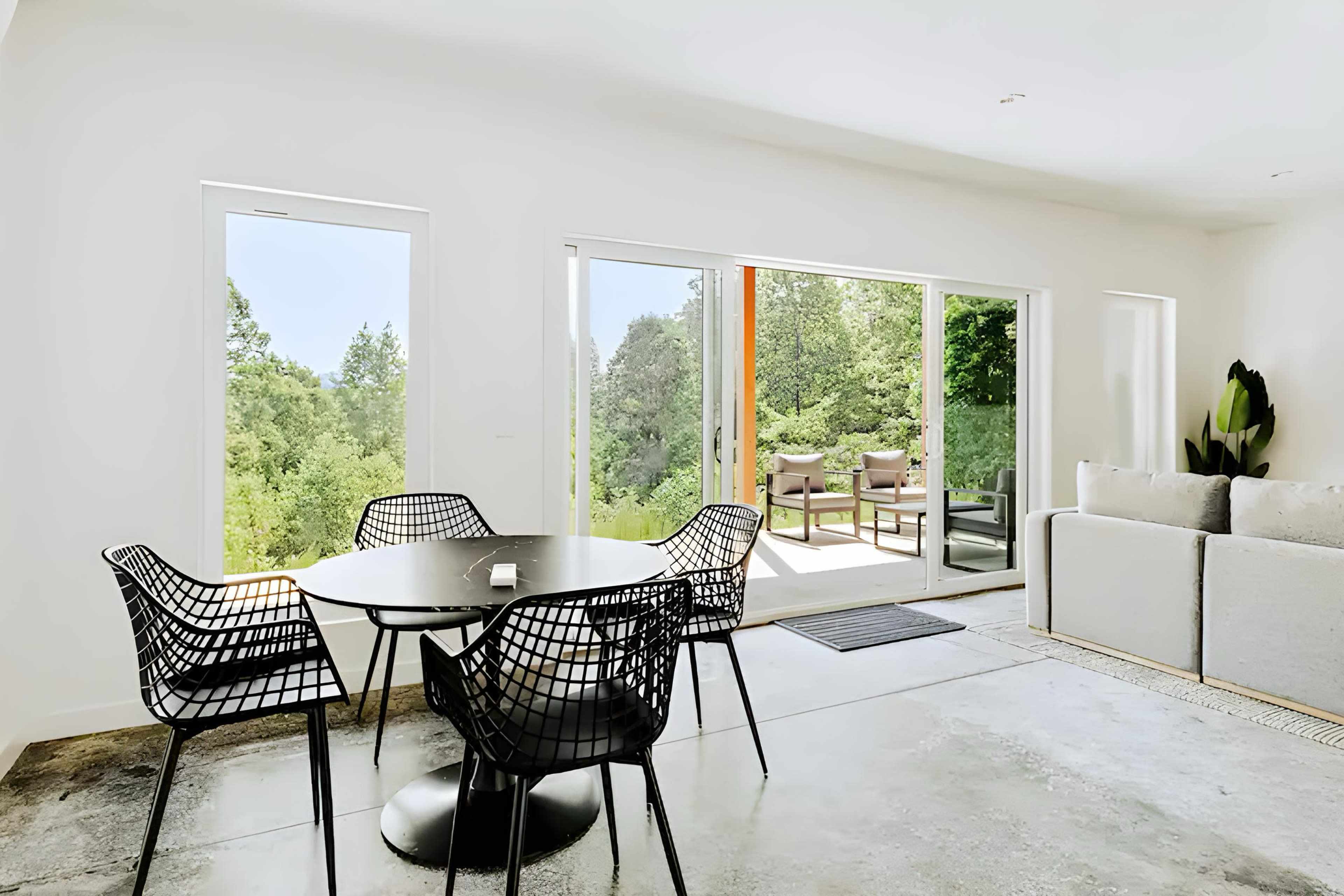 A modern dining area features a round table surrounded by six black wire chairs, with large sliding doors leading to a view of greenery outside.