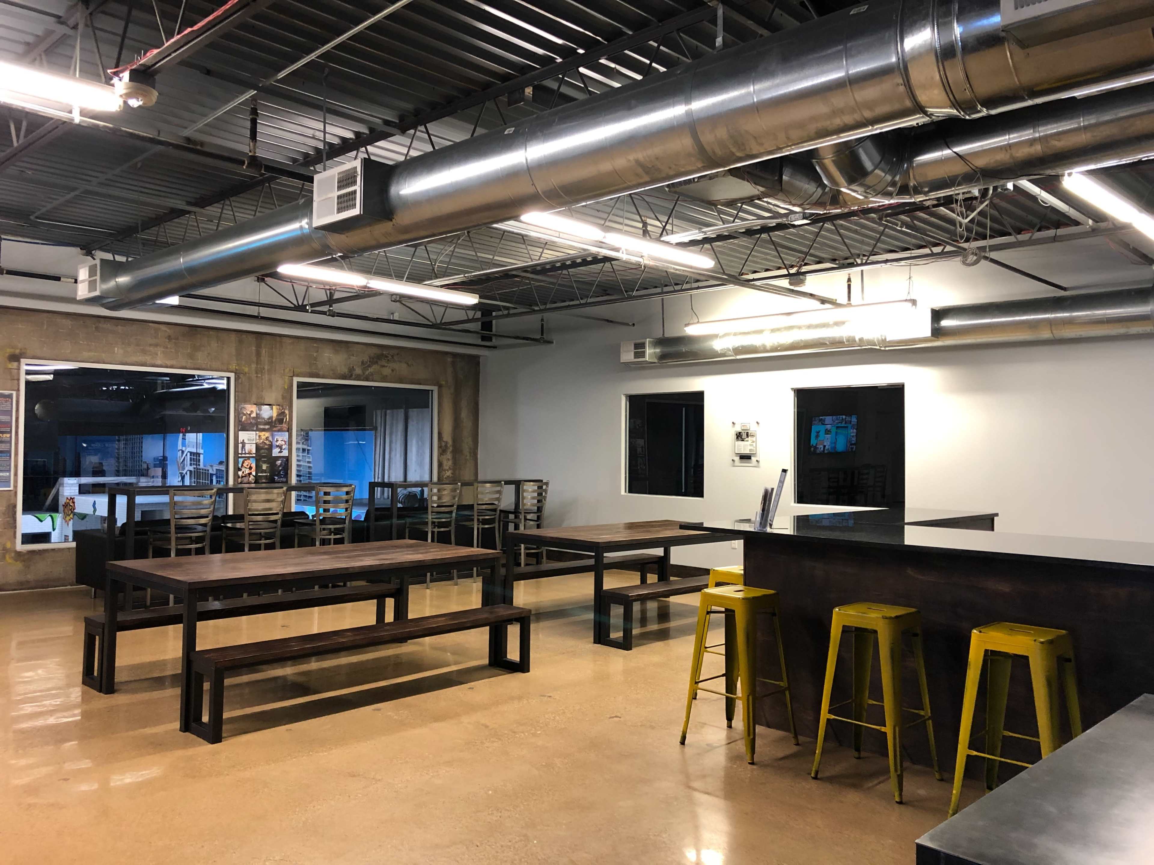 A spacious industrial-style workspace with wooden tables, metal bar stools, and exposed ductwork on the ceiling.