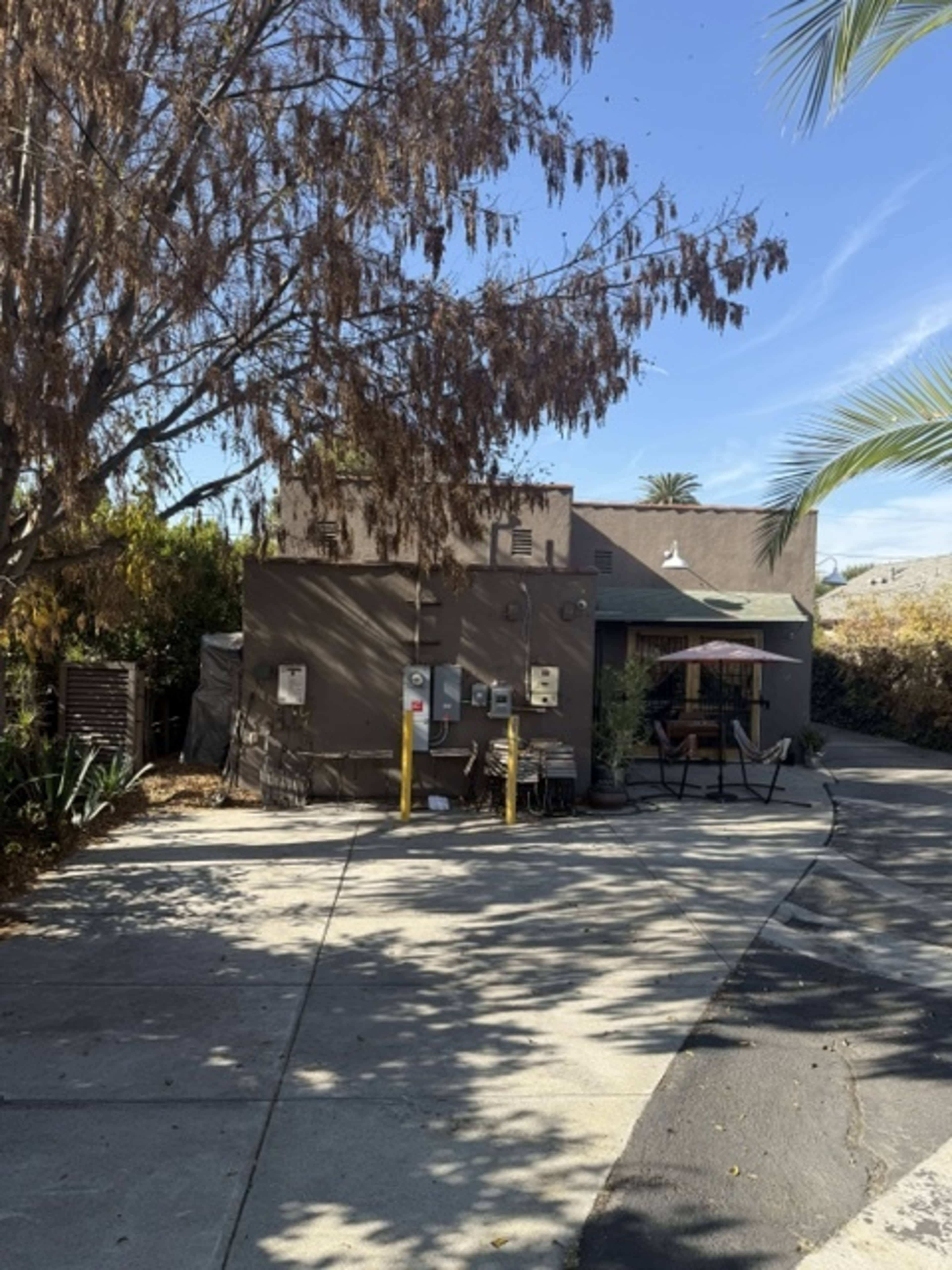 A narrow driveway leads to a brown building partially shaded by a tree, with outdoor furniture visible nearby.