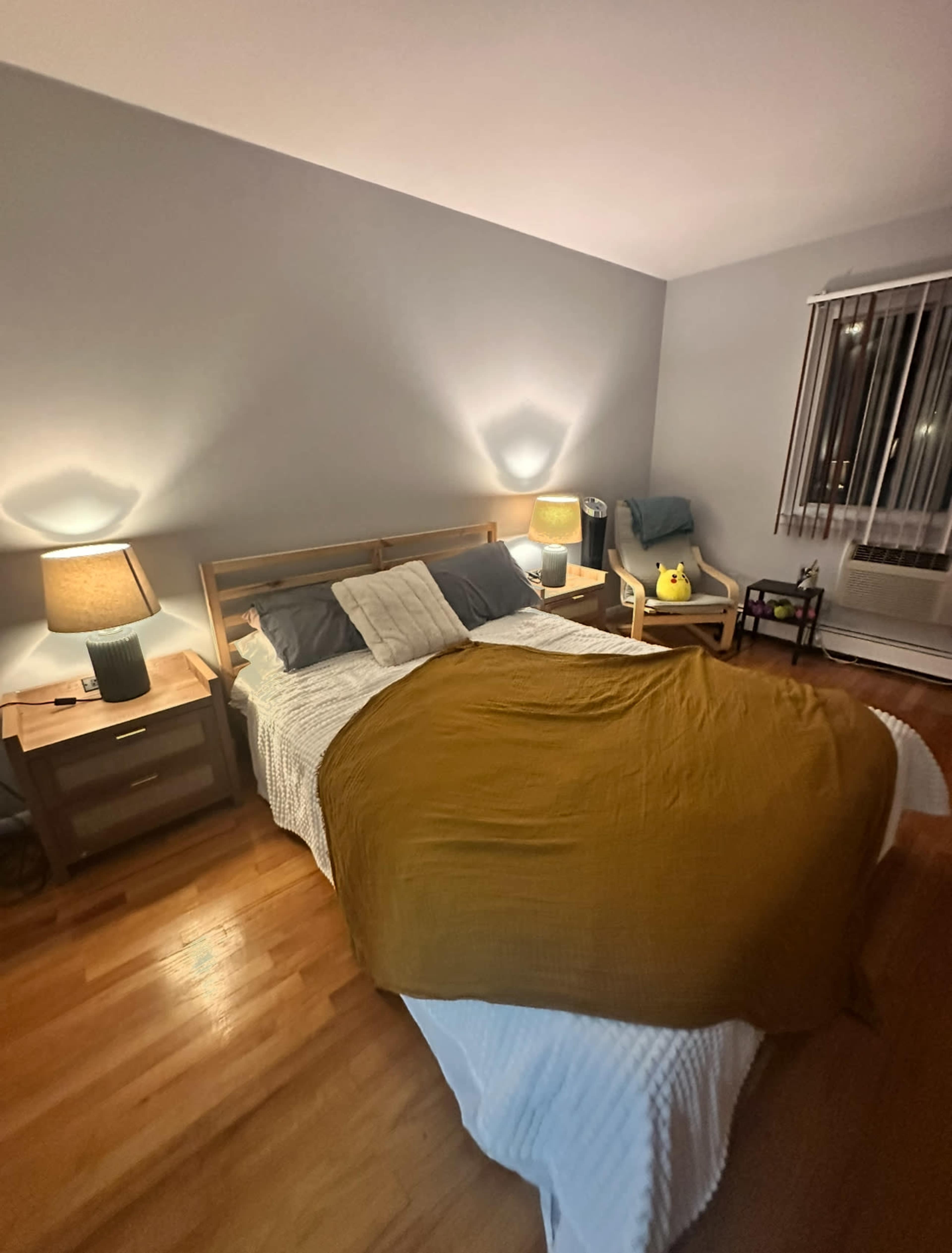 A neatly made bed with a mustard-colored blanket is positioned in a softly lit bedroom featuring wooden furniture and blinds.