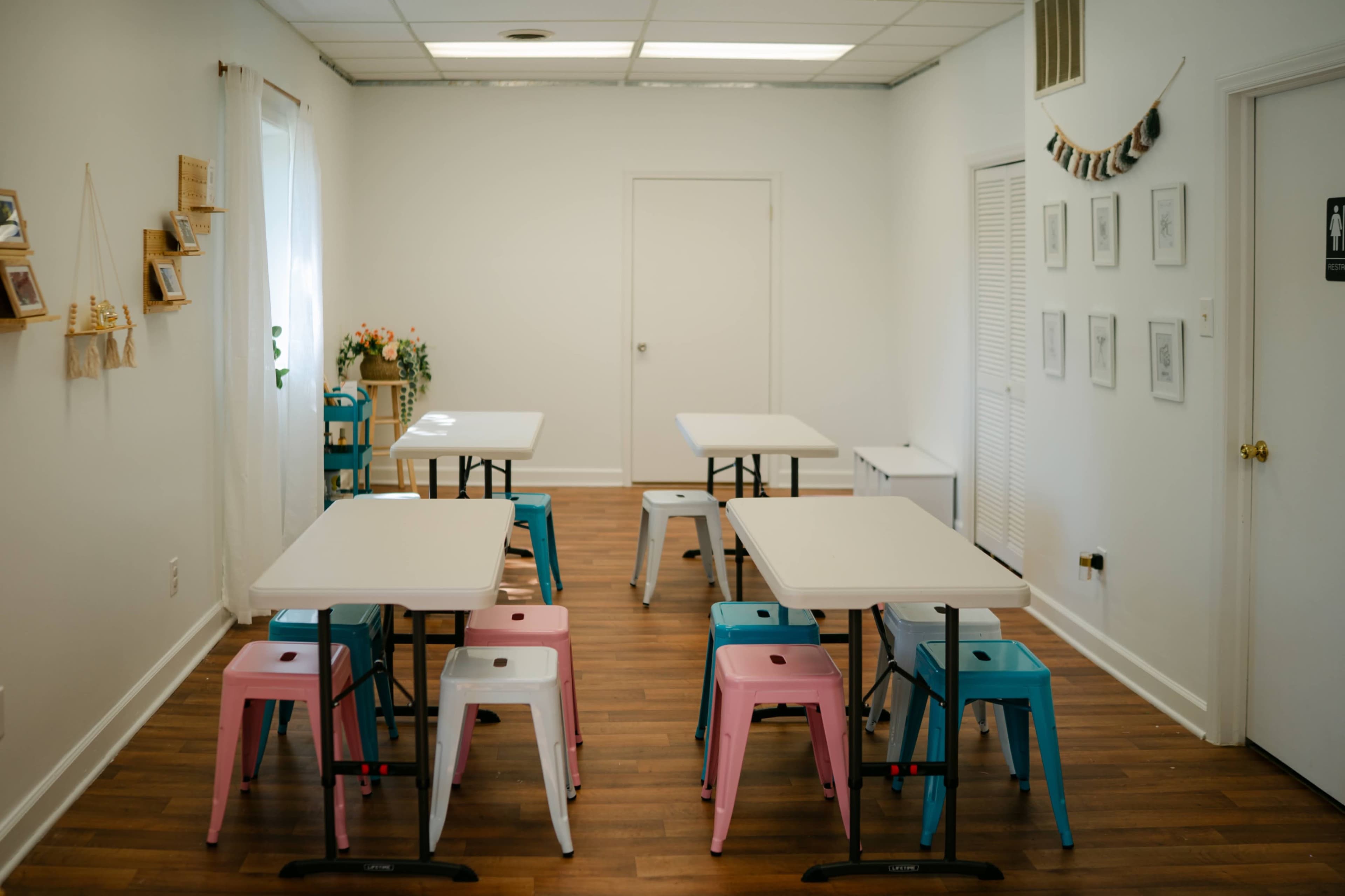 The image shows a bright, minimalist room with several tables and colorful stools arranged neatly.