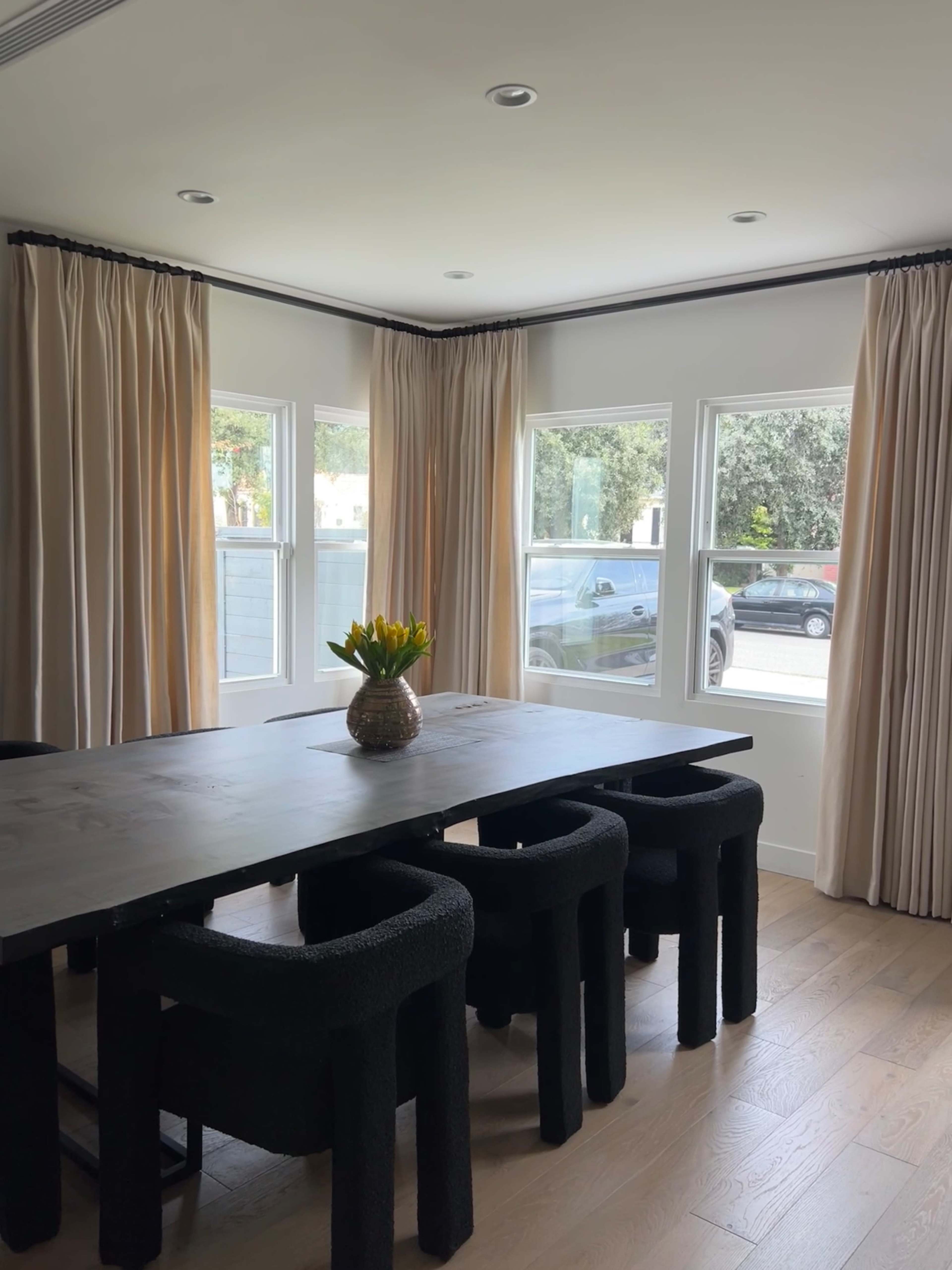 A dining area features a large wooden table surrounded by black upholstered chairs and a vase of yellow flowers on the table, with light-colored curtains framing the windows.