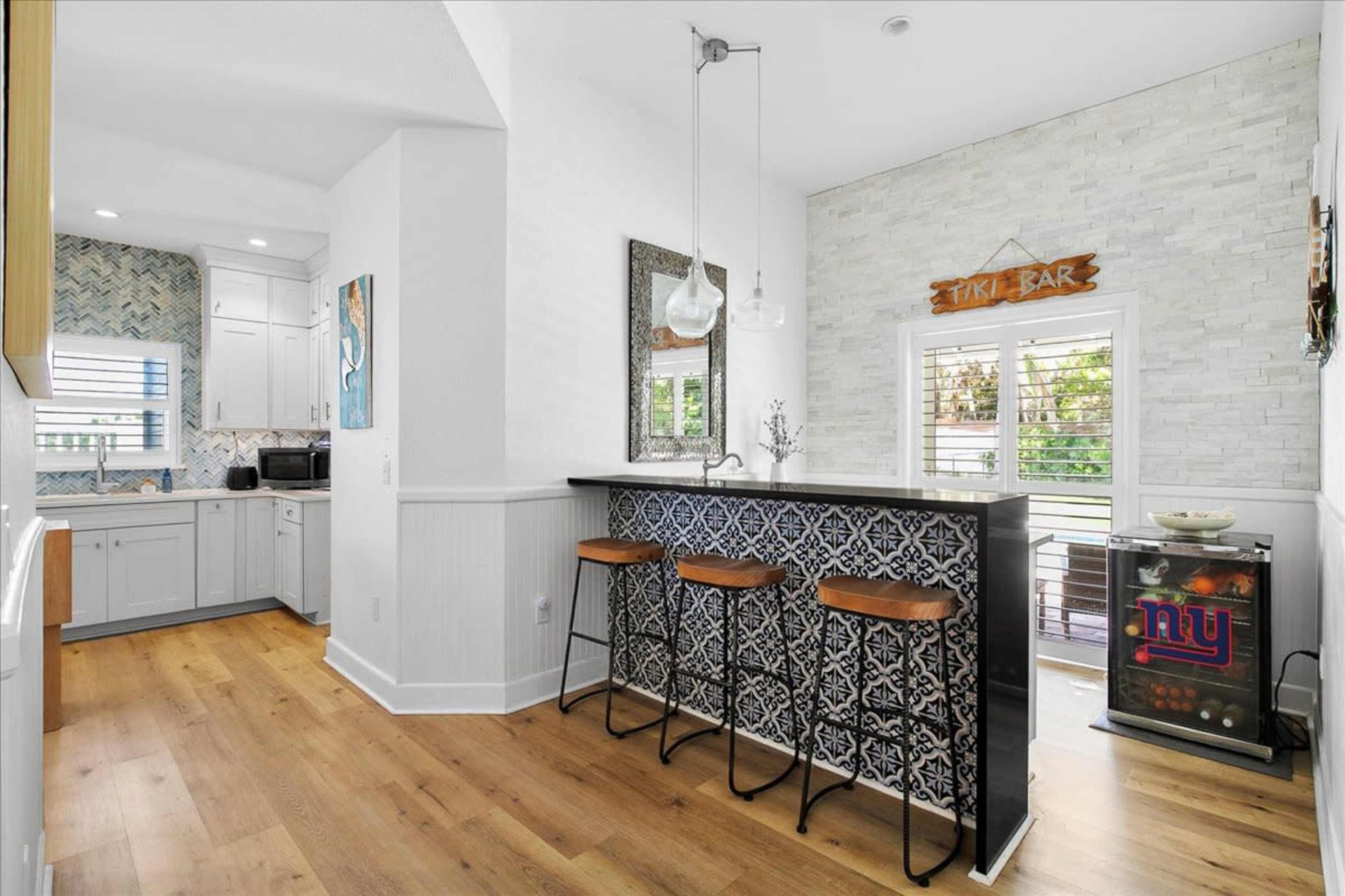 A modern kitchen features a bar area with three stools, a patterned backsplash, and a view of a small outdoor space.