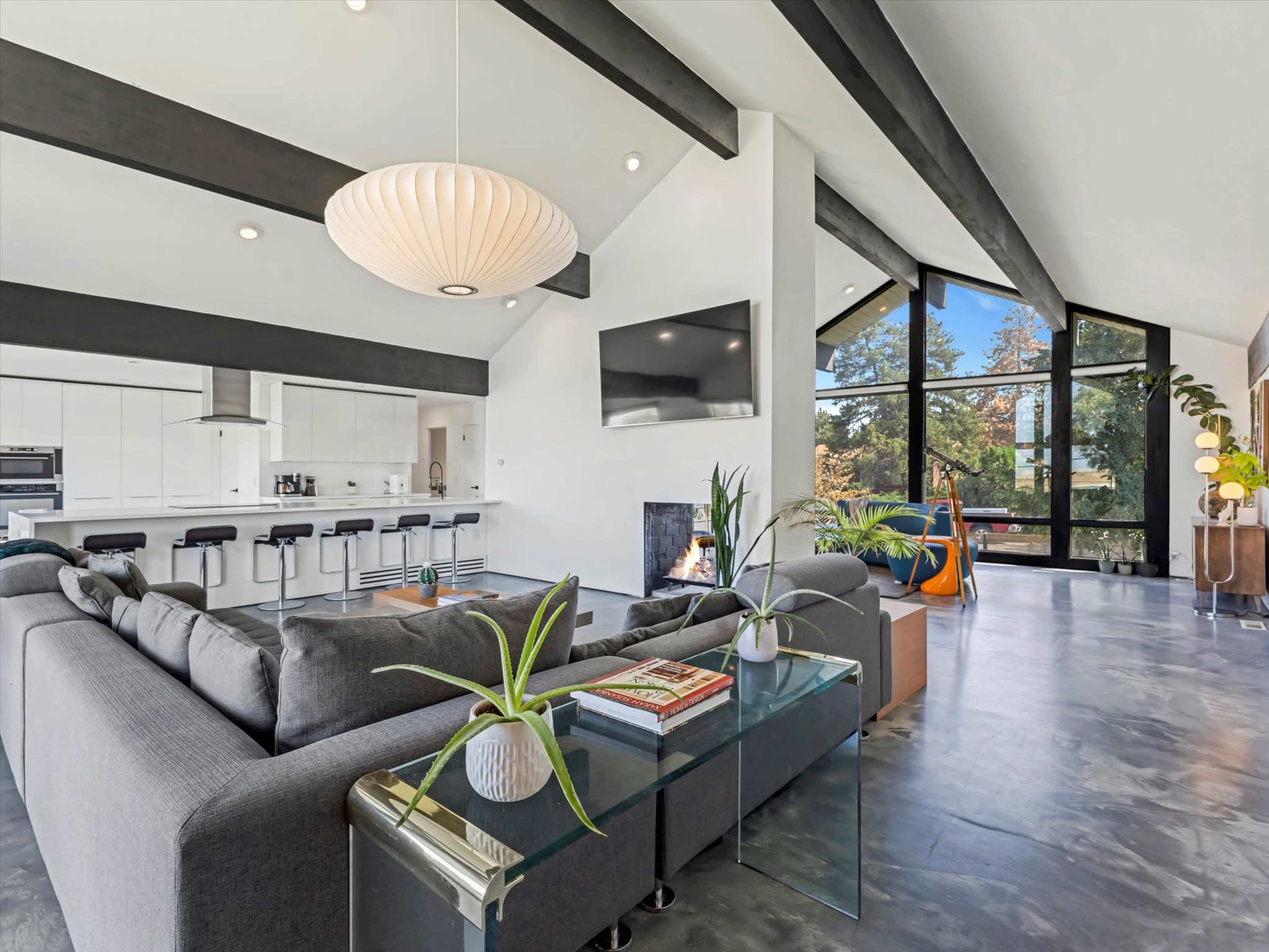 The image shows a modern living room with a gray sectional sofa, a wall-mounted television, and a large window that provides natural light.