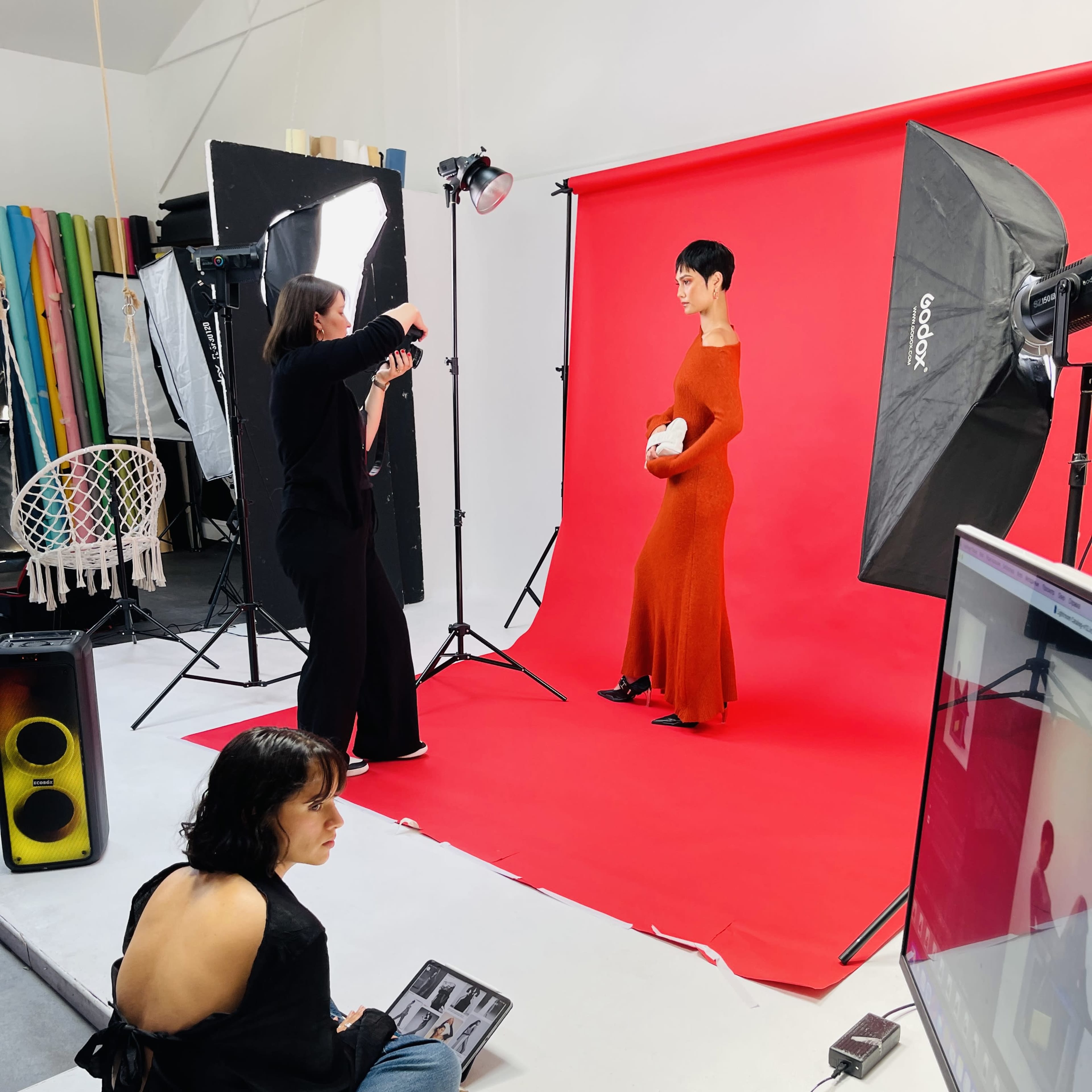 A model poses in a long red dress against a bright red backdrop while a photographer captures the shot, and a stylist reviews looks on a laptop.
