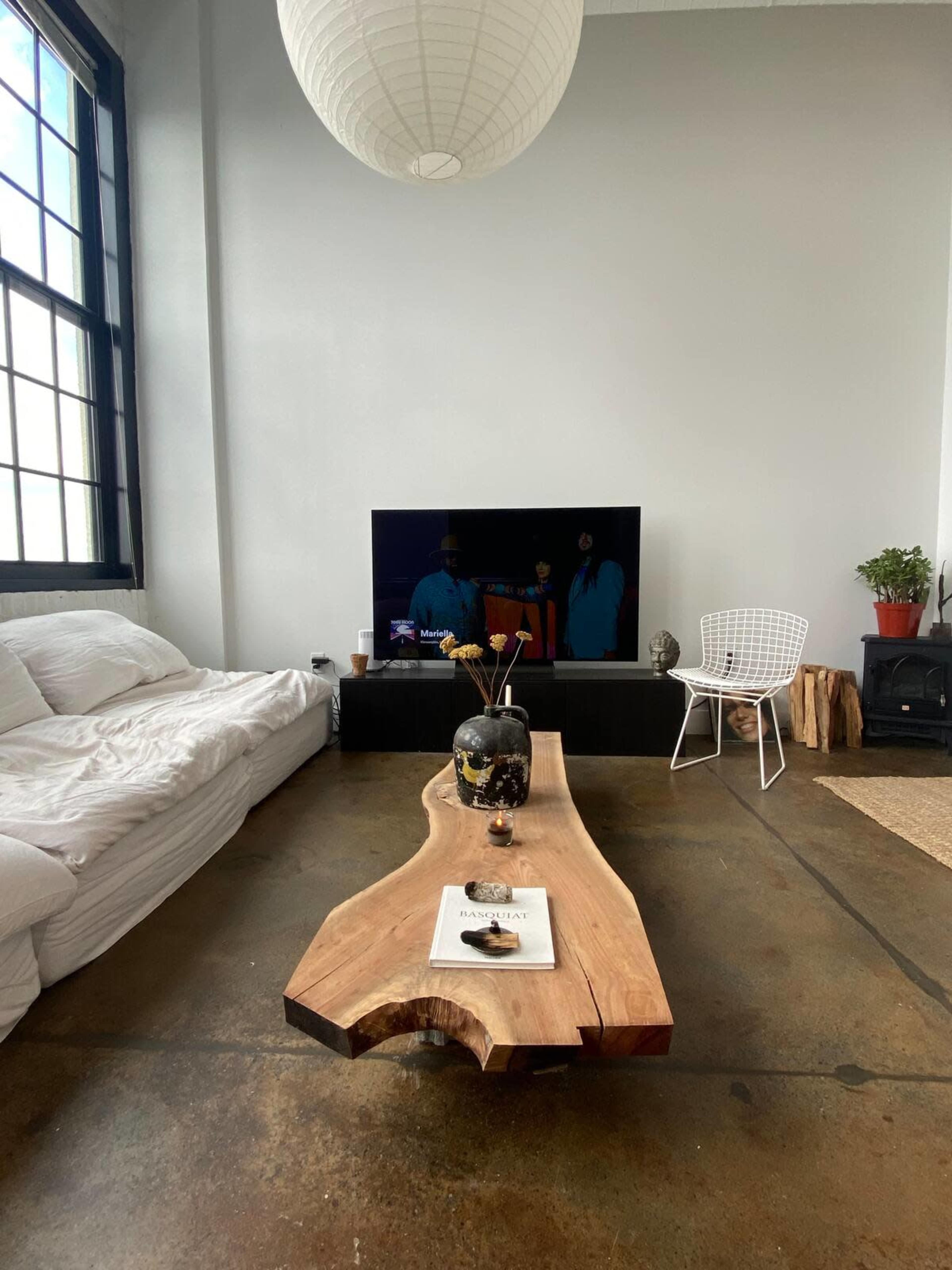 A spacious living room features a large coffee table made of wood, a white sofa, a television on a dark cabinet, and a potted plant near a window.