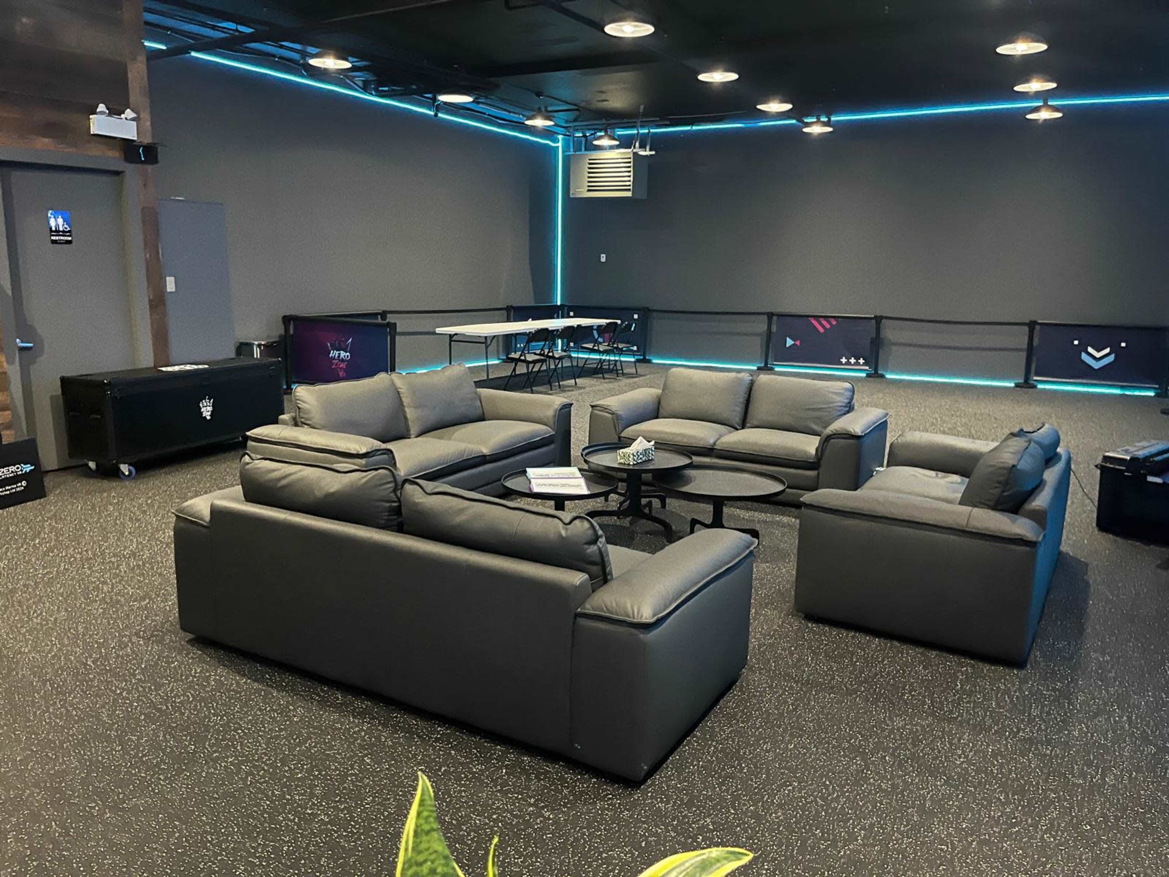 The image shows a modern lounge area with gray sectional sofas arranged around a circular coffee table in a spacious room with dark walls and blue LED lighting.