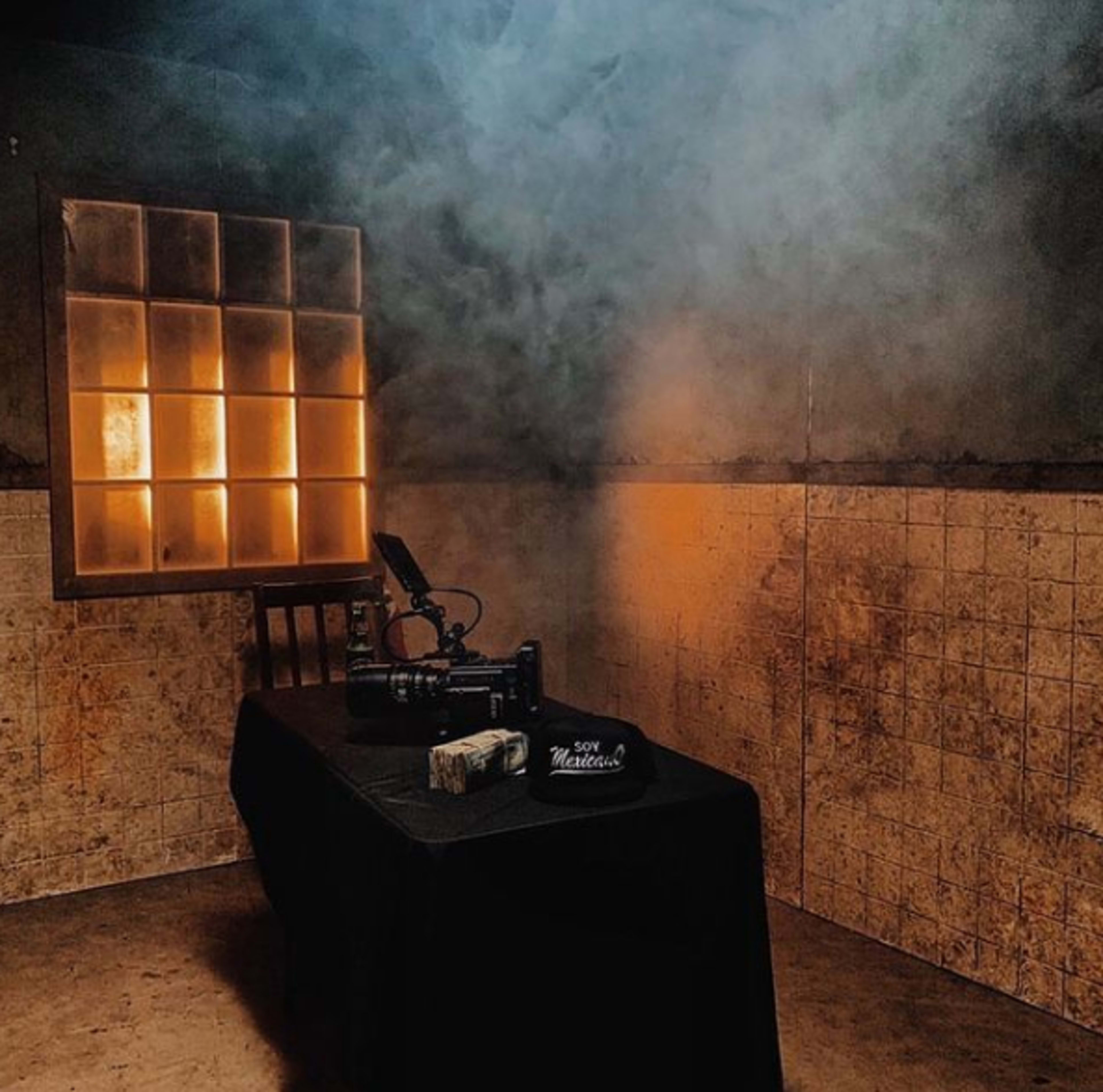 A camera sits on a table covered with a black cloth in a dimly lit, smoky room with a window casting an orange glow.