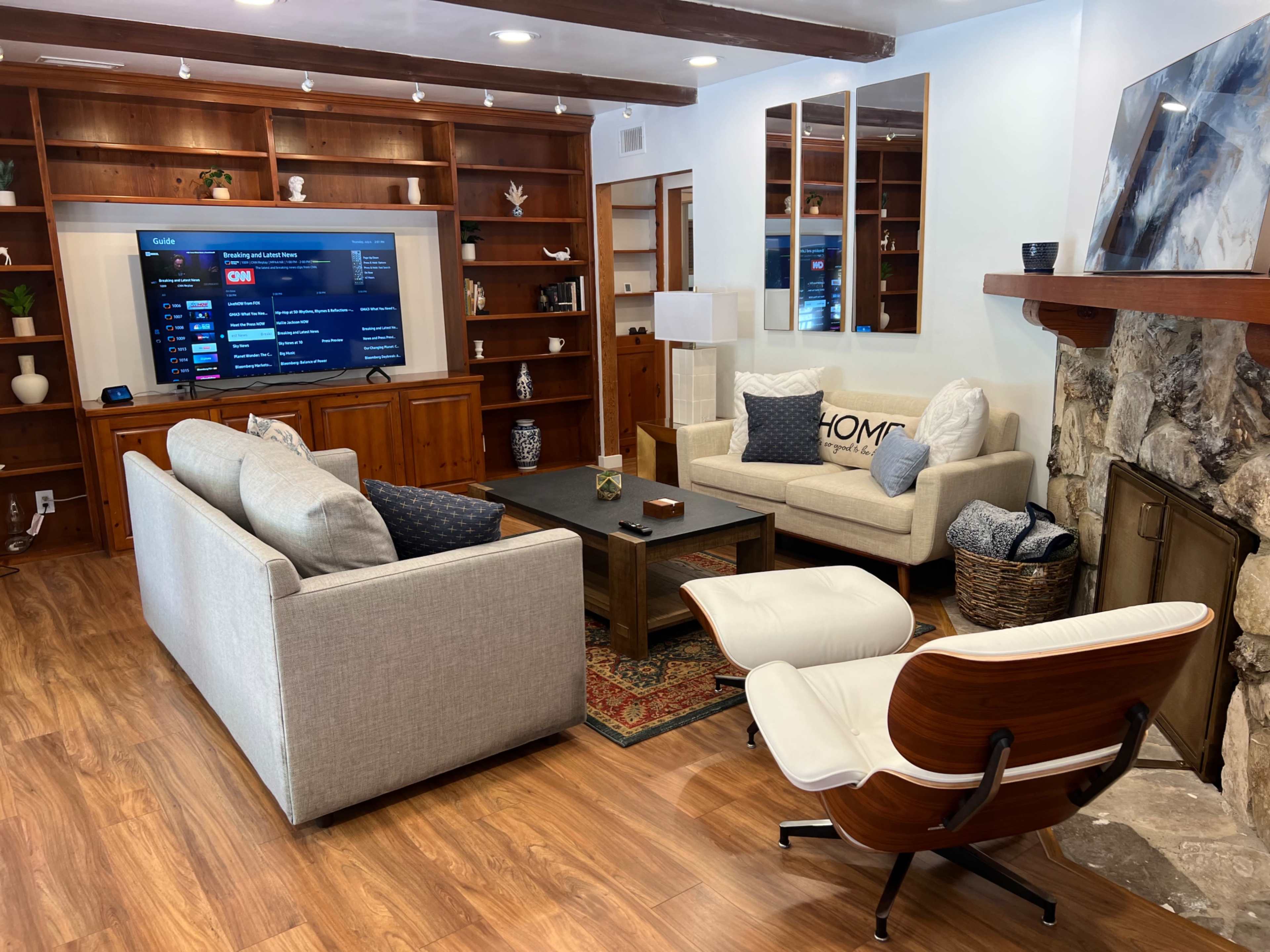 A living room features two sofas, a coffee table, a television mounted on the wall, and a stone fireplace, with wooden shelves and a rug enhancing the space.