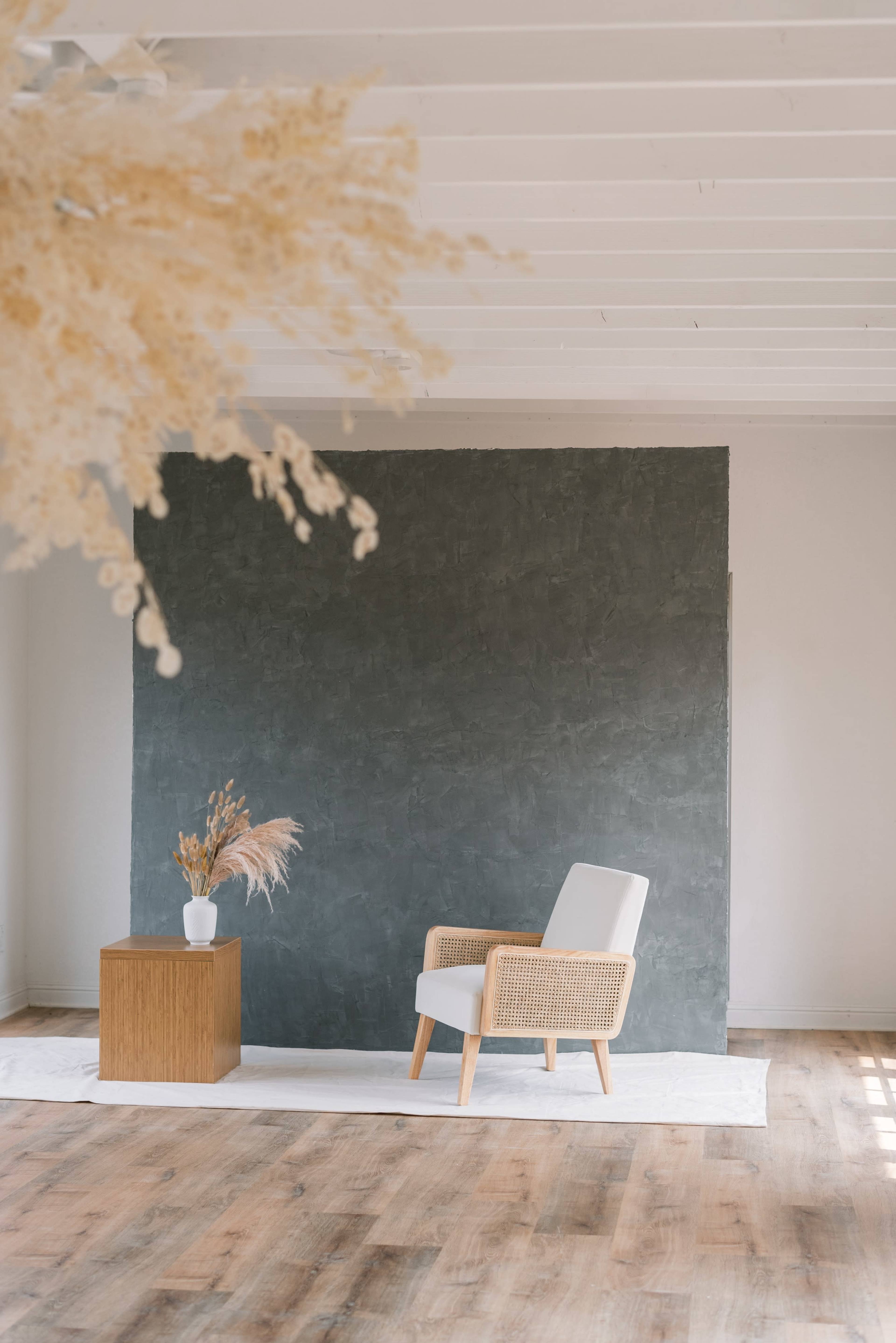 A minimalist interior scene features a rattan chair beside a wooden table, with a vase of dried flowers against a dark textured wall.
