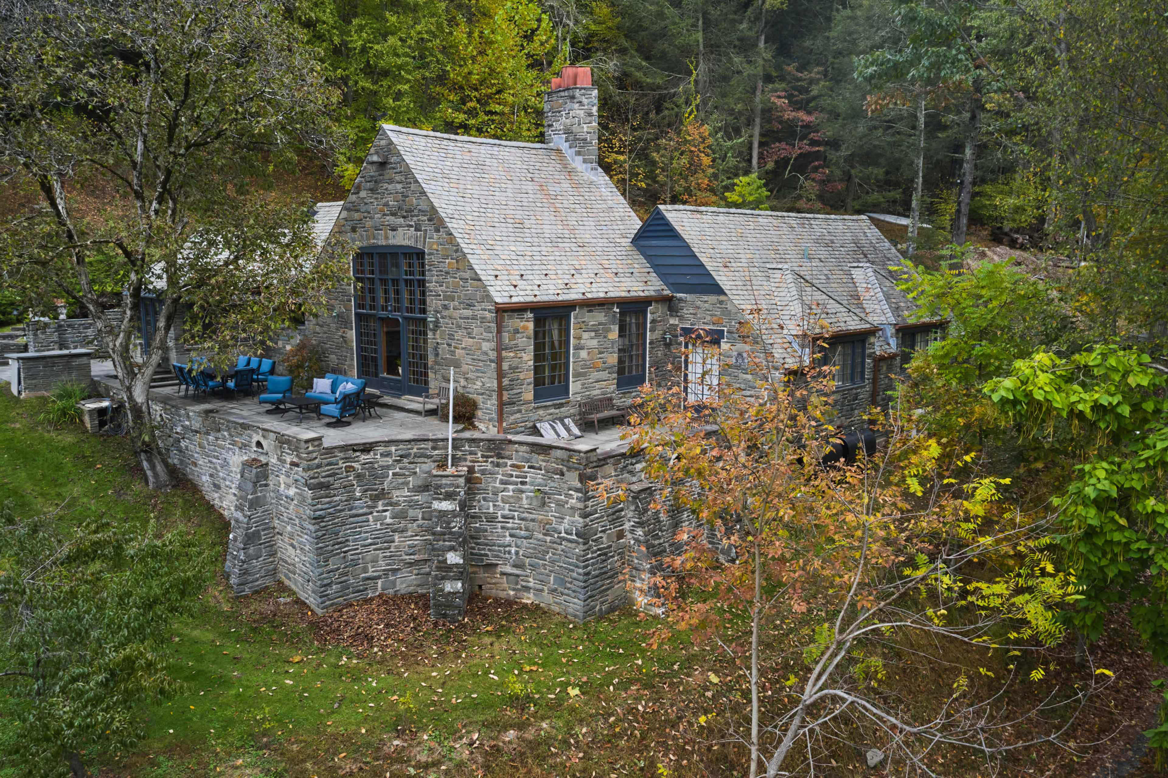 The image shows a stone cottage surrounded by trees, featuring a large terrace with blue chairs and a chimney.