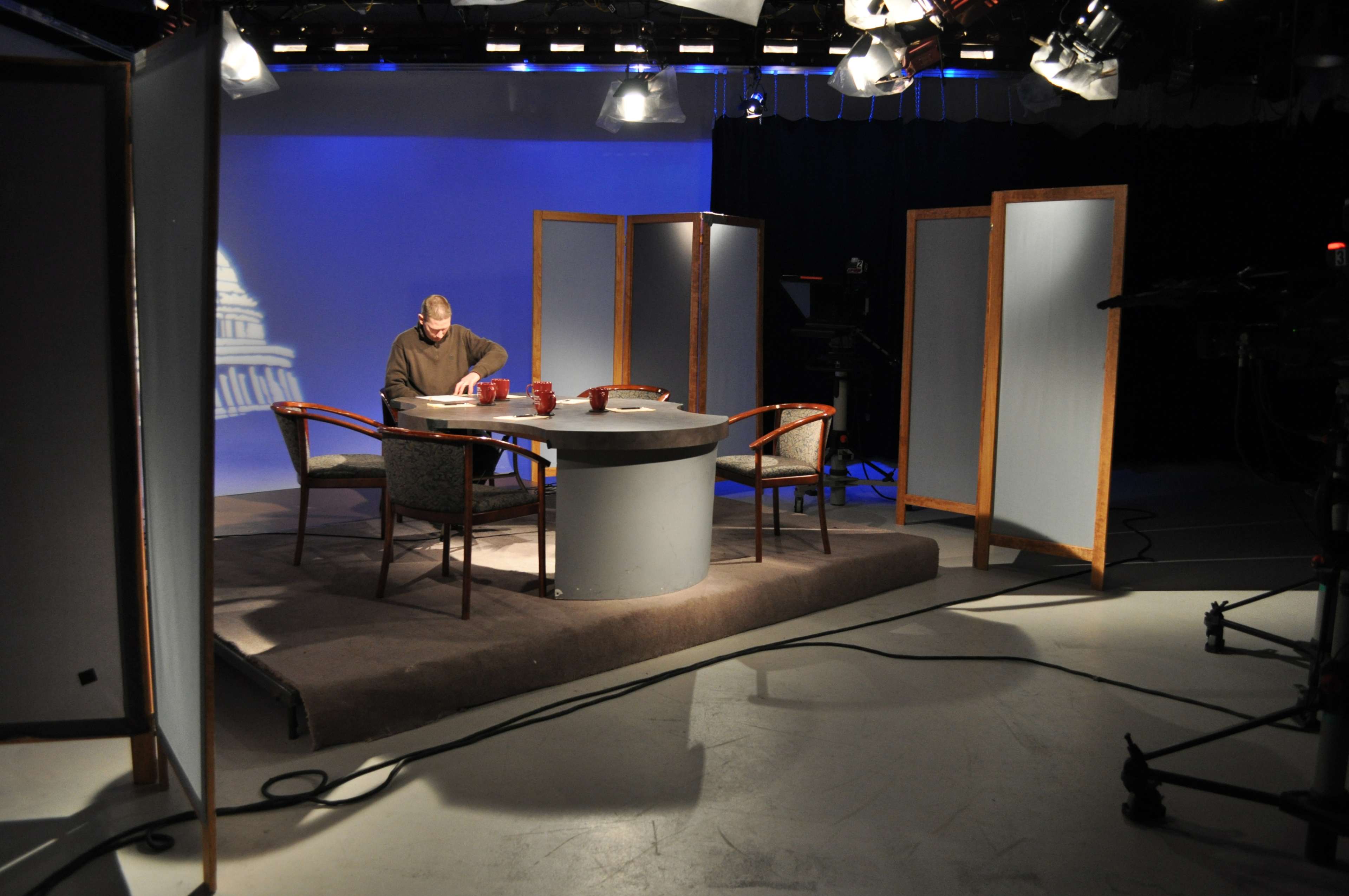 A television studio setup features a circular table with four chairs, a man sitting at the table, and backdrop screens with a faint image of a building.