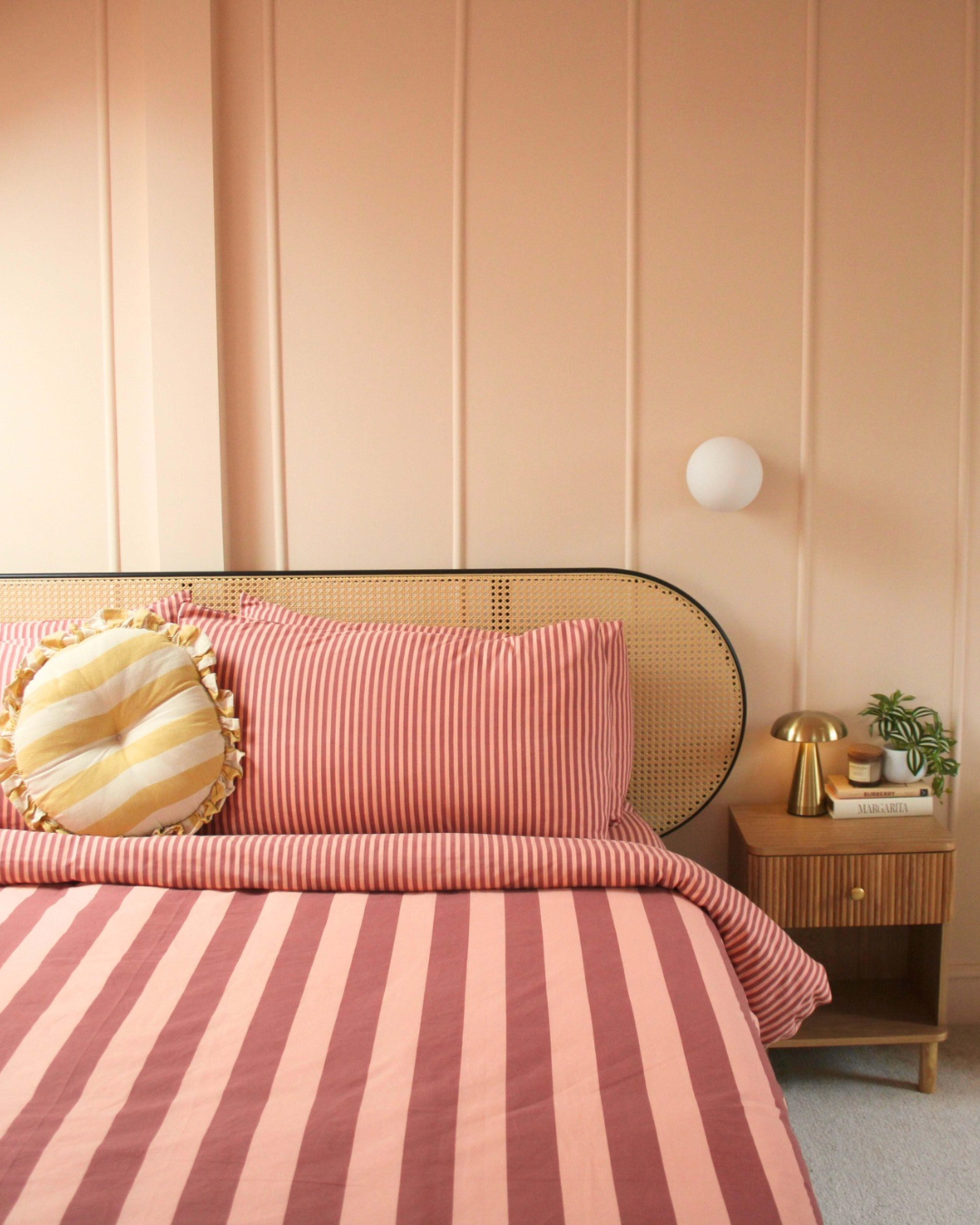 The image depicts a neatly made bed with striped pink and coral bedding, a round decorative pillow, and a stylish bedside table with a lamp and a small plant.