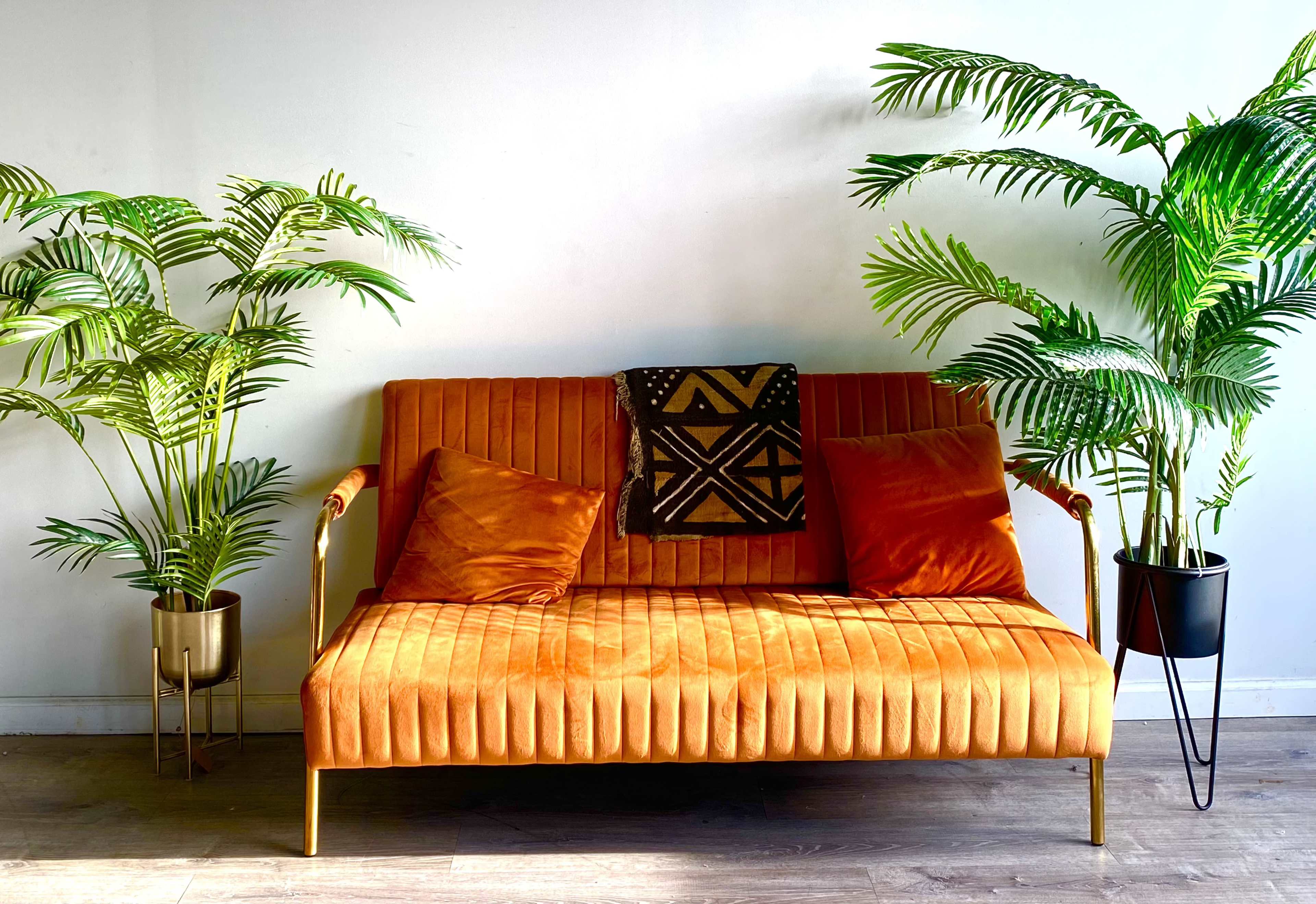 A velvet orange loveseat is flanked by two tall potted plants against a white wall.