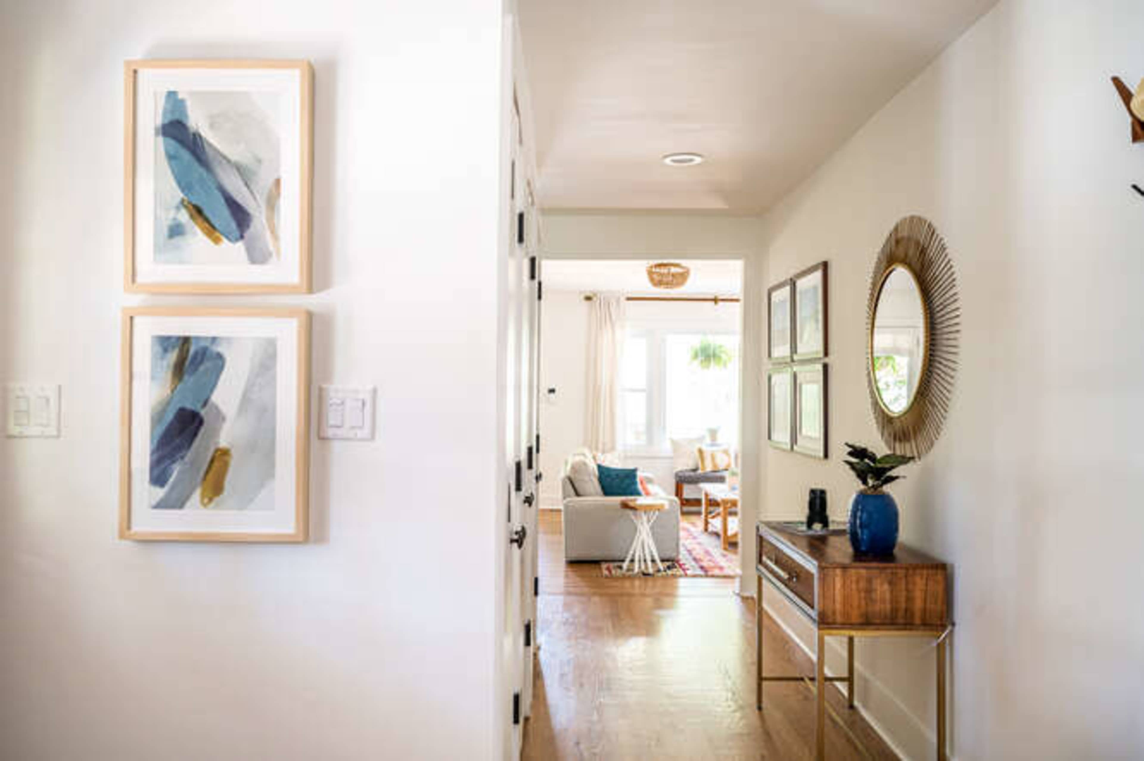 A hallway features two framed artworks on the wall beside a console table with a plant and a round mirror, leading to a living room area.