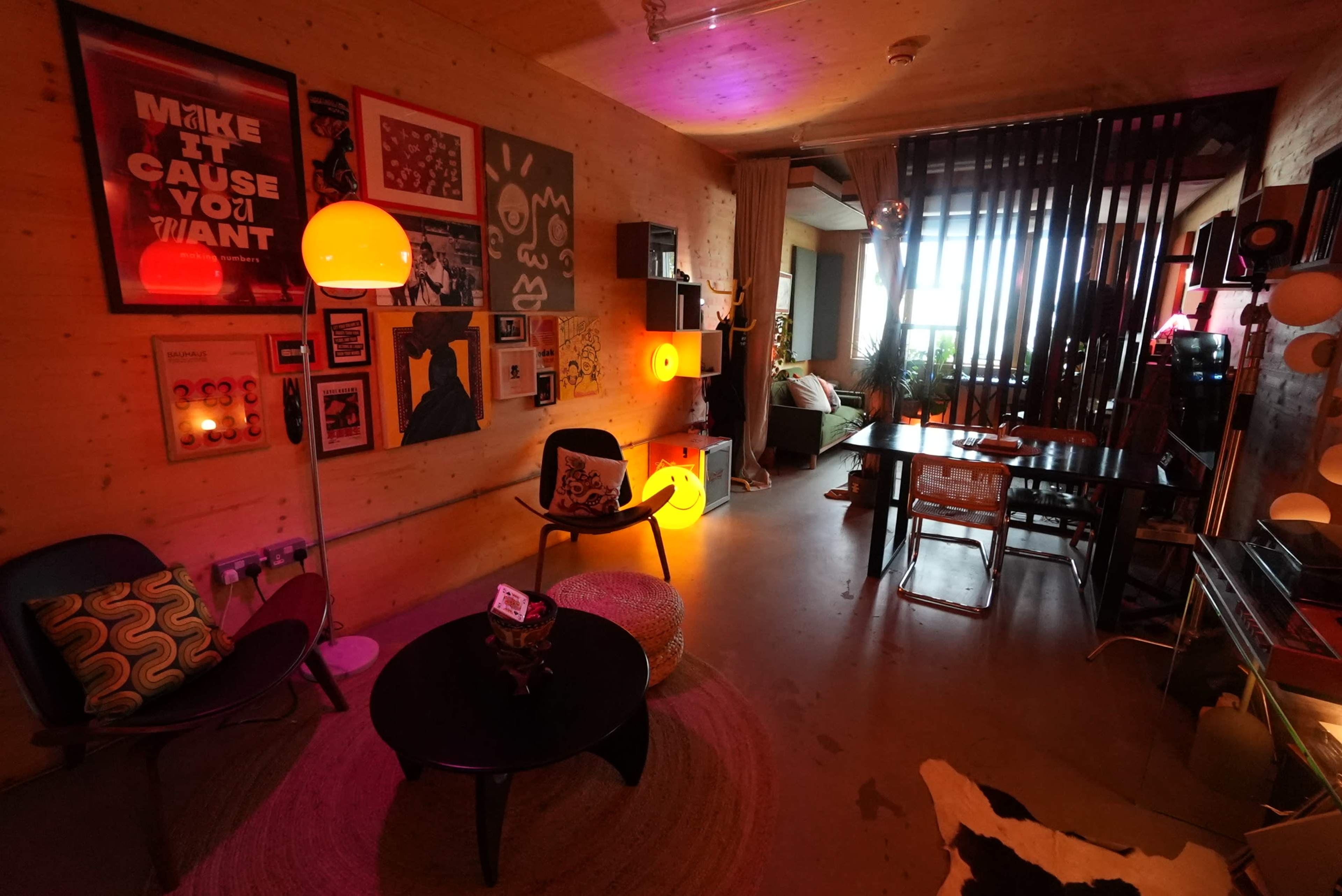 The image shows a stylish interior space featuring modern furniture, decorative artwork on the walls, and various light sources creating a vibrant atmosphere.