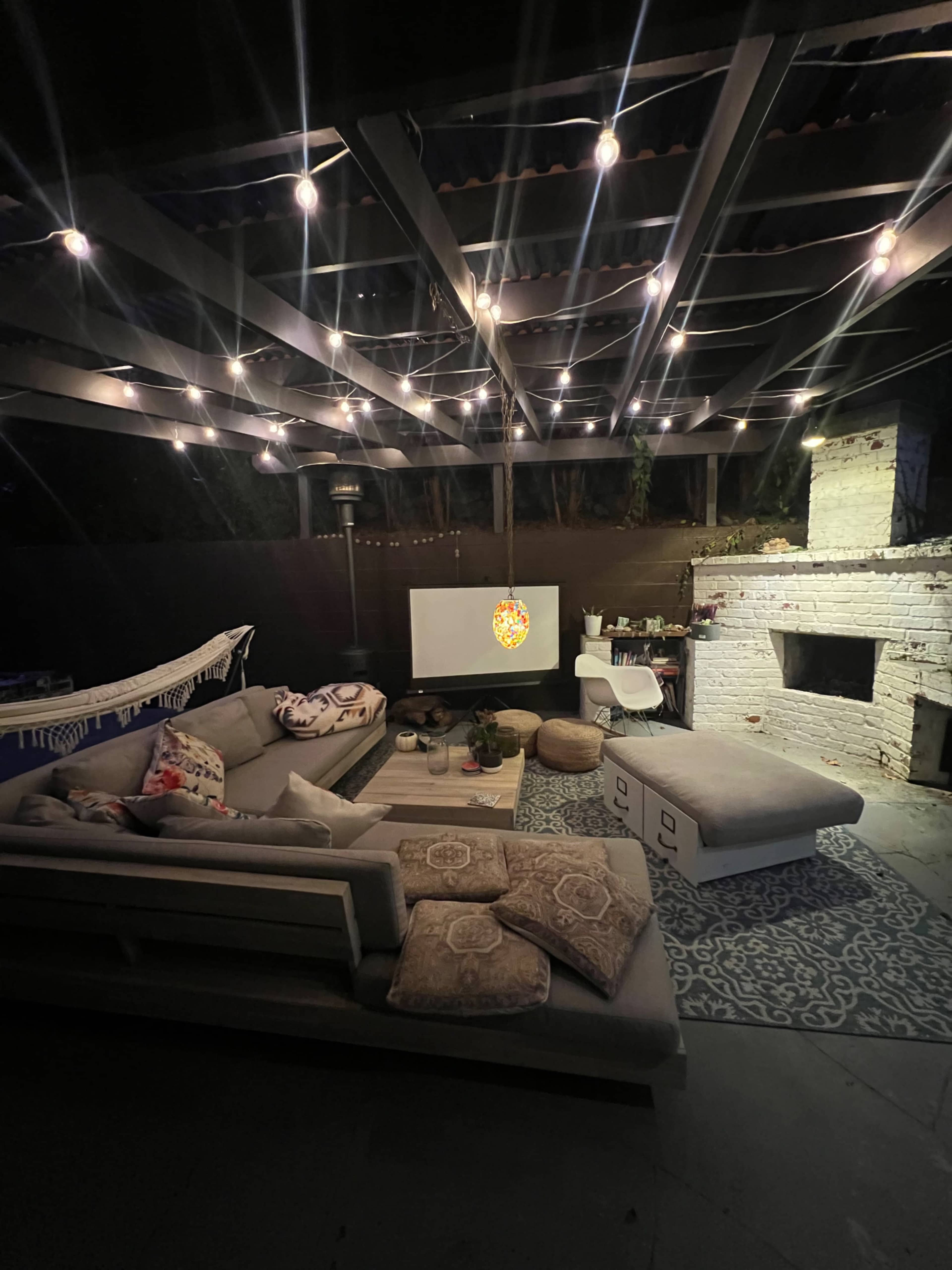 A cozy outdoor living area features a sectional sofa, a large TV screen, and string lights overhead, set against a backdrop of a white brick wall.