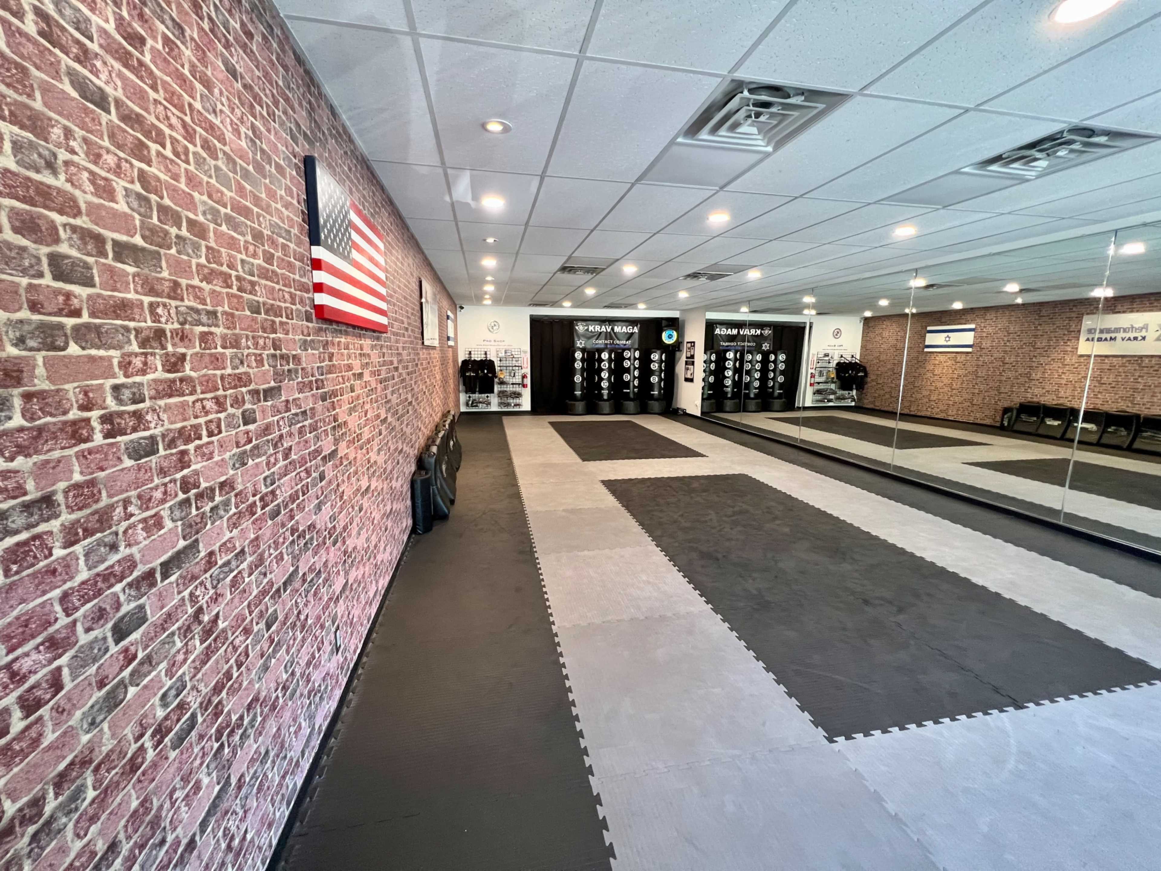 A martial arts training room featuring a brick-patterned wall, black and gray mats, an American flag, and mirrors.