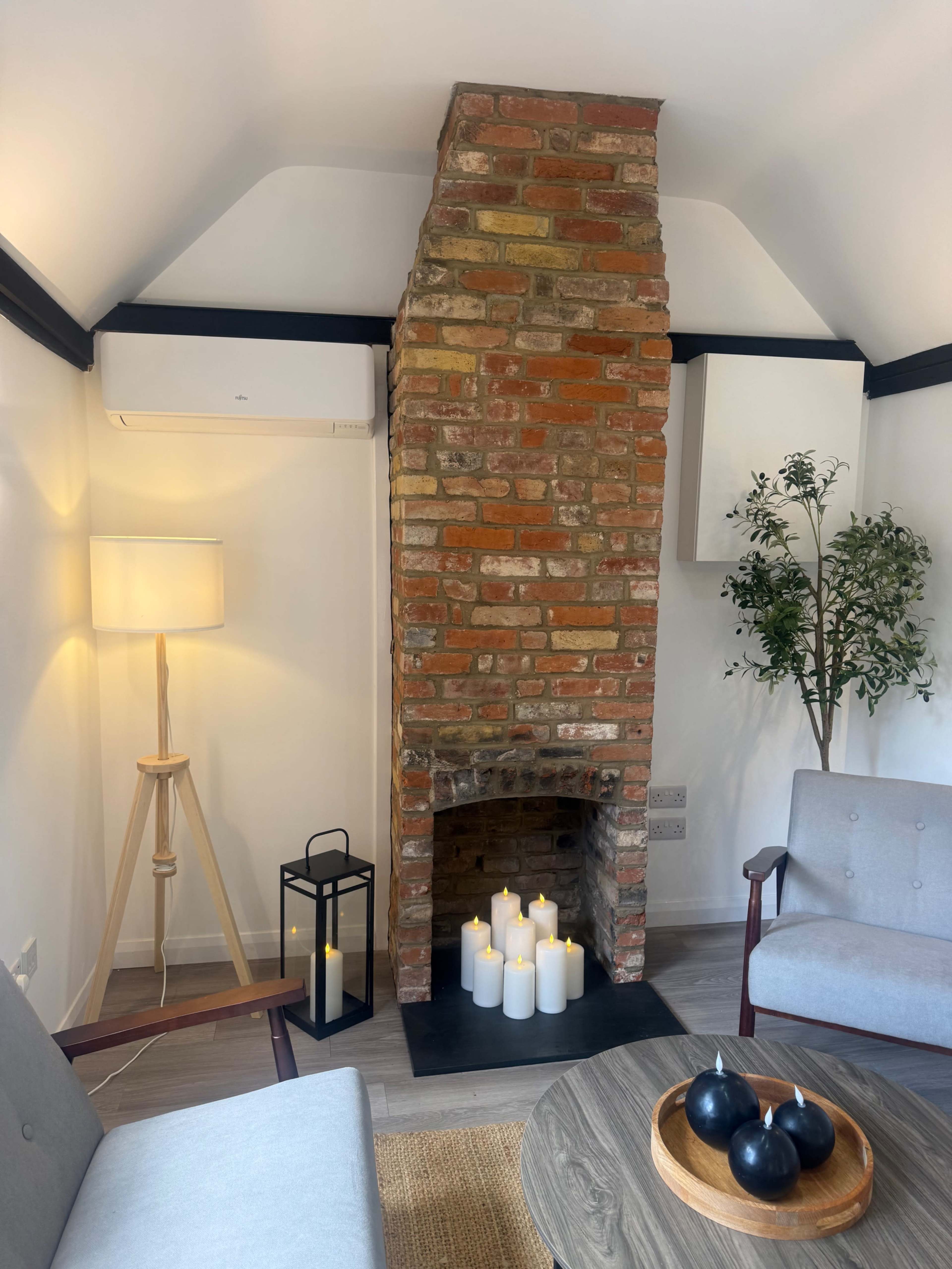 The image shows a cozy living space featuring a brick fireplace with candles, a round wooden table, and two gray armchairs, accompanied by a floor lamp and a small indoor plant.