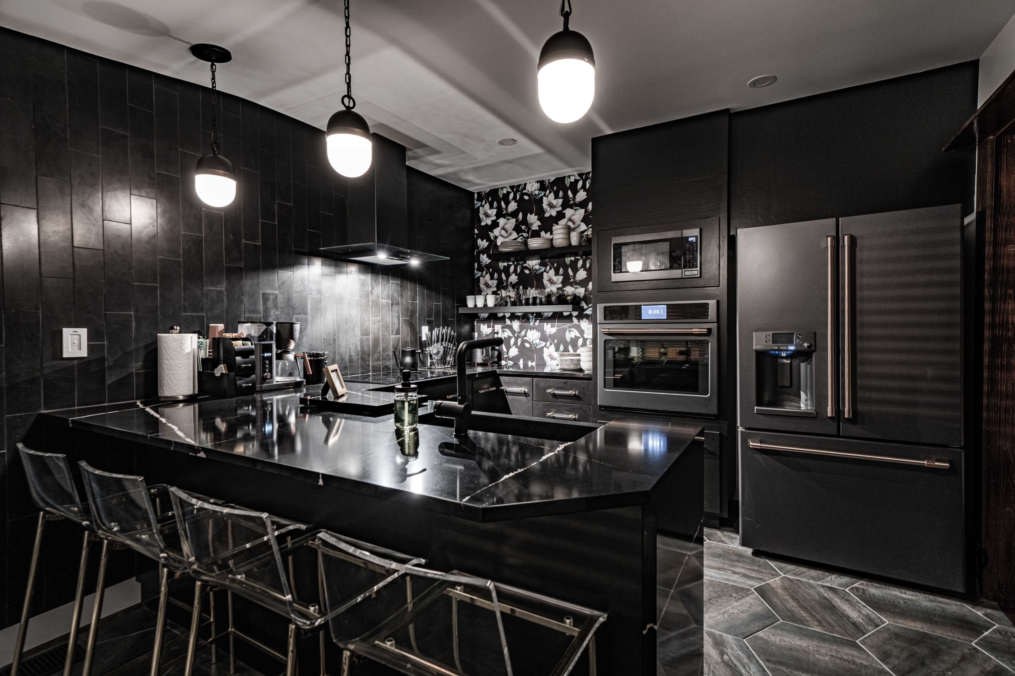 A modern kitchen features dark cabinetry, sleek appliances, and a black island with transparent bar stools under pendant lighting.