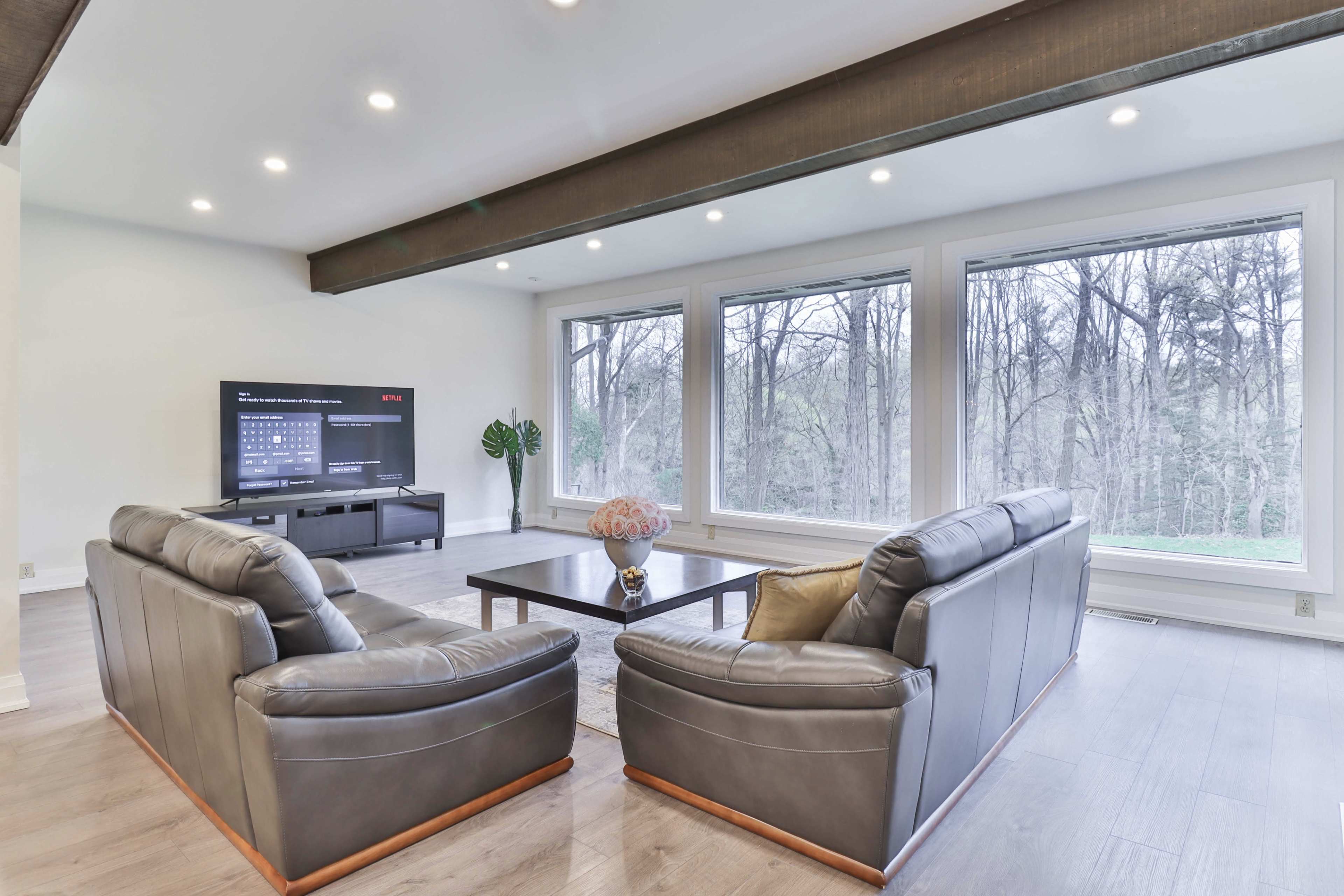 The scene features a modern living room with two leather sofas facing a coffee table, a television mounted on the wall, and large windows overlooking a wooded area.